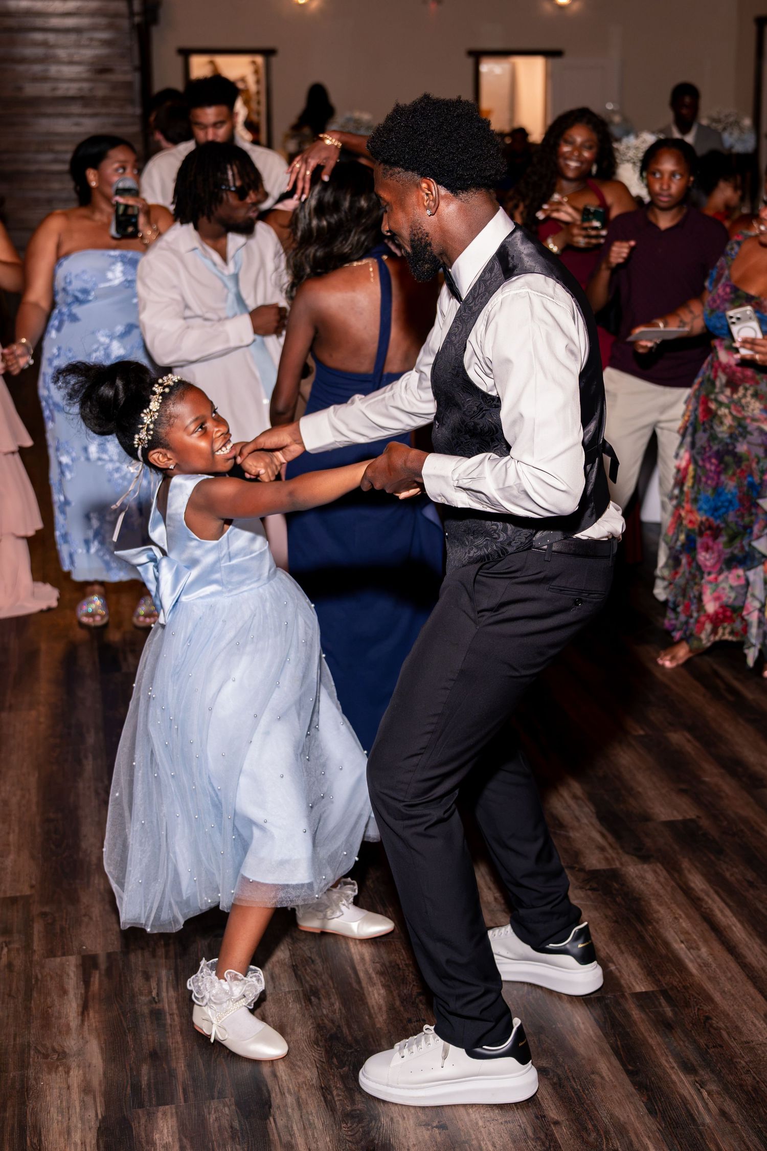Groom in white shirt dances playfully with young flower girl in blue dress at wedding.