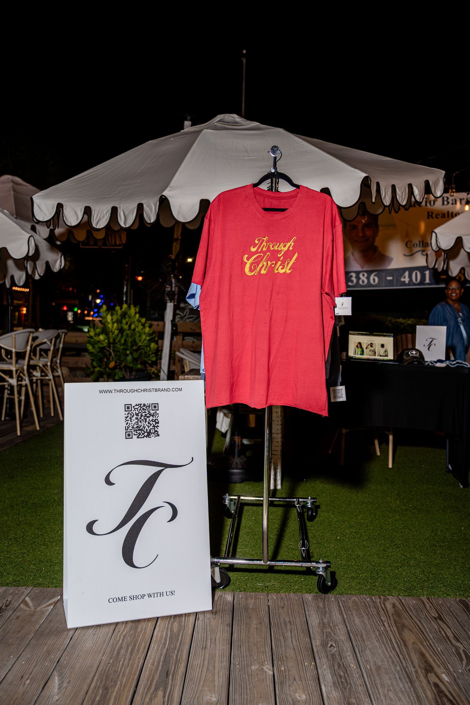 Red Through Christ graphic t-shirt displayed on metal rack with TC brand QR code sign outdoors.