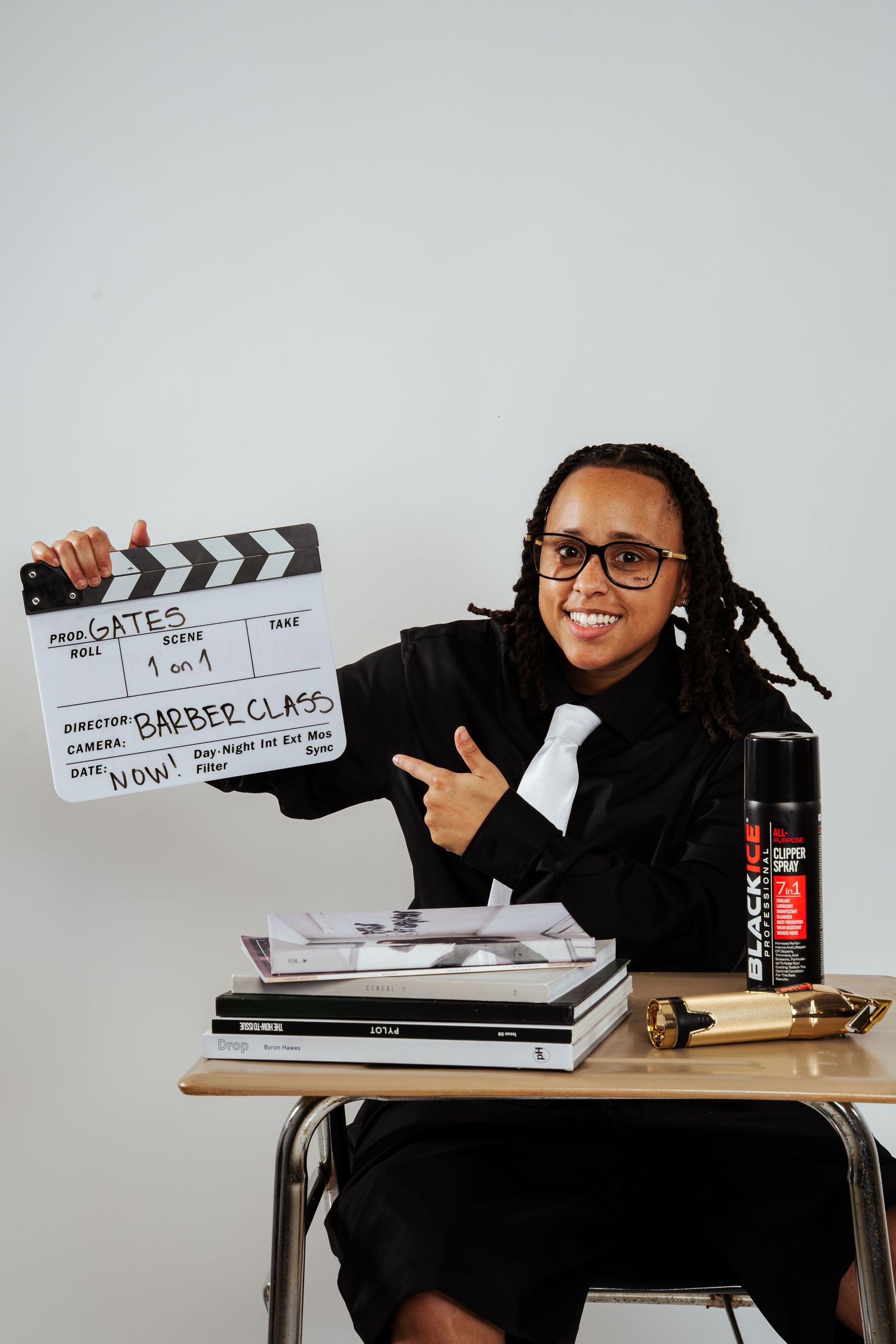 Smiling person in black suit pointing at Barber Class film clapperboard seated at school desk with products.