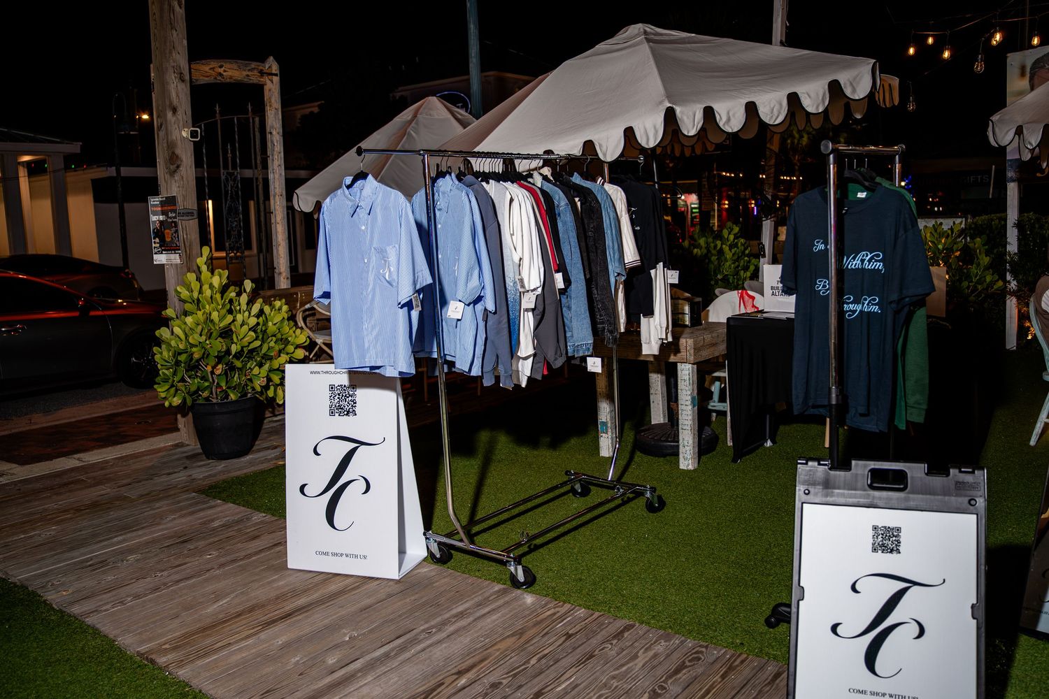 Outdoor clothing rack display with TC brand shirts under white umbrella at night market event.
