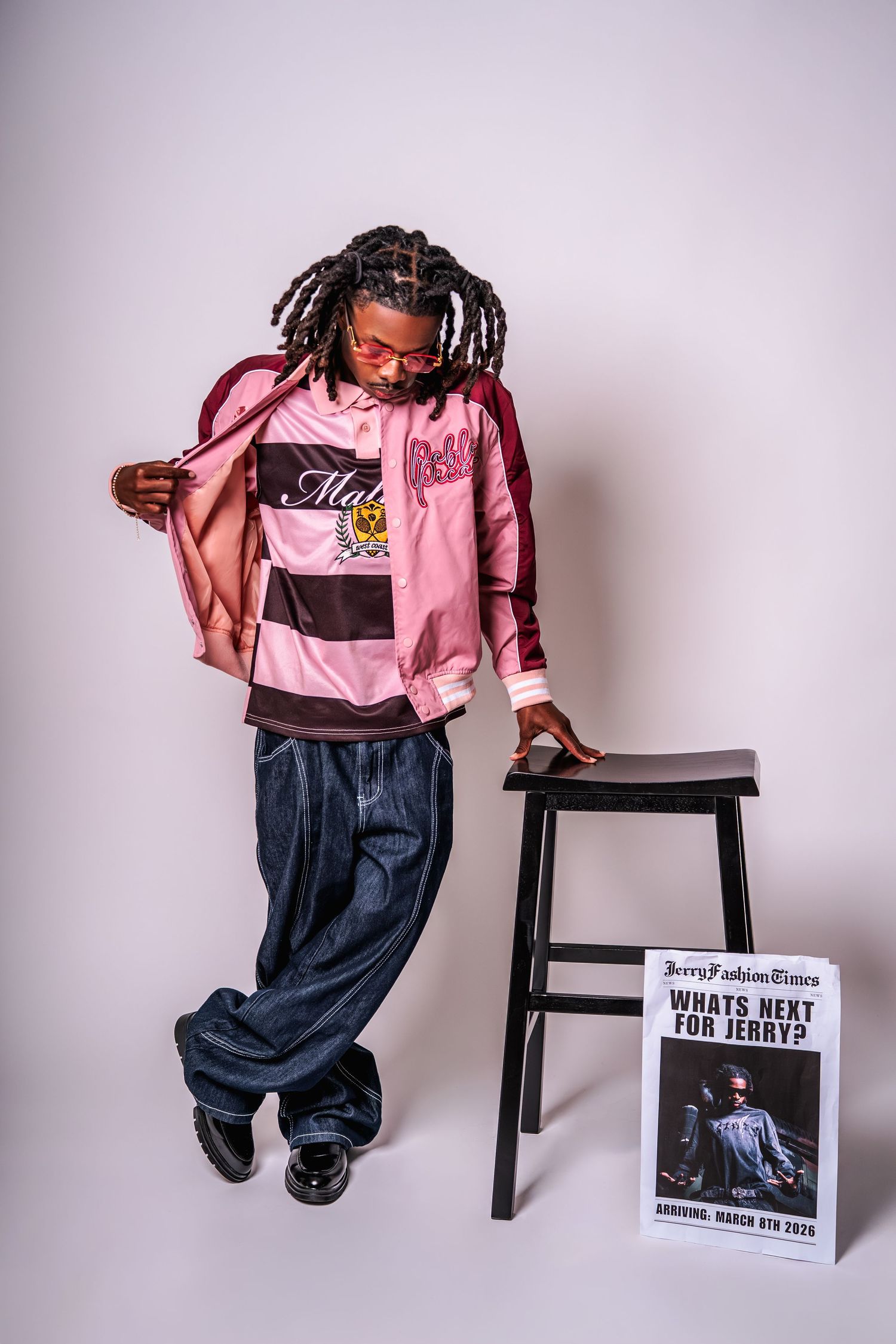 Stylish young man with dreadlocks posing in a pink varsity jacket and wide-leg jeans next to a promotional sign.