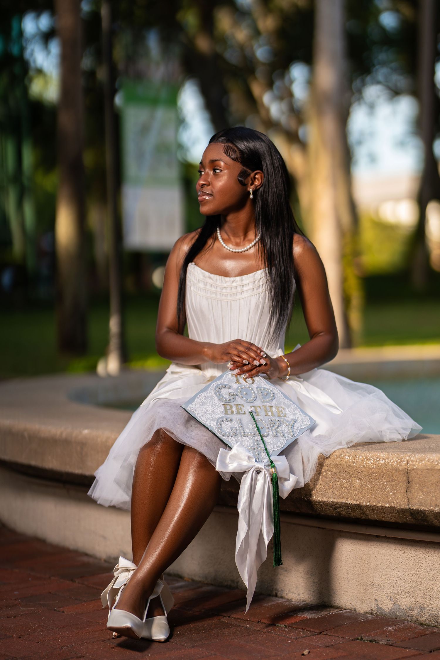 Young graduate in white dress holds decorated cap while seated outdoors among trees at golden hour.