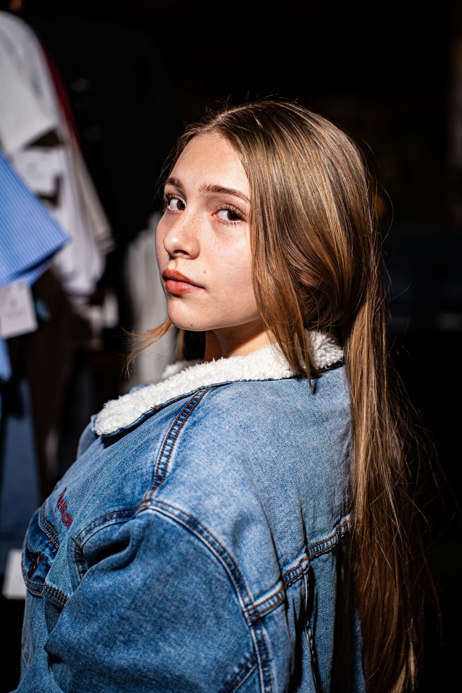 Teen girl in denim sherpa jacket posing at night with clothing rack in background.