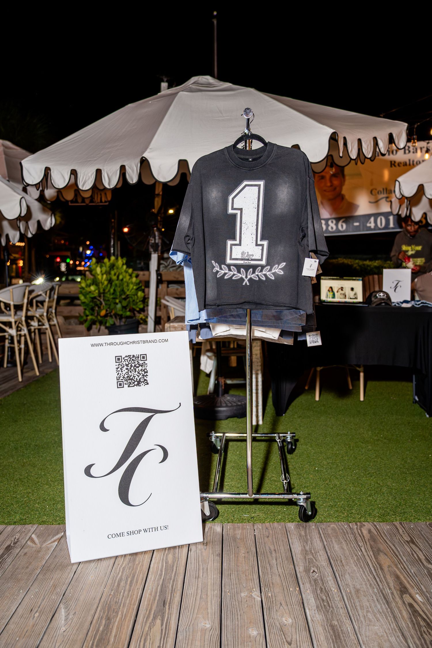 Black number one graphic t-shirt displayed on rack with TC brand sign at outdoor night event.