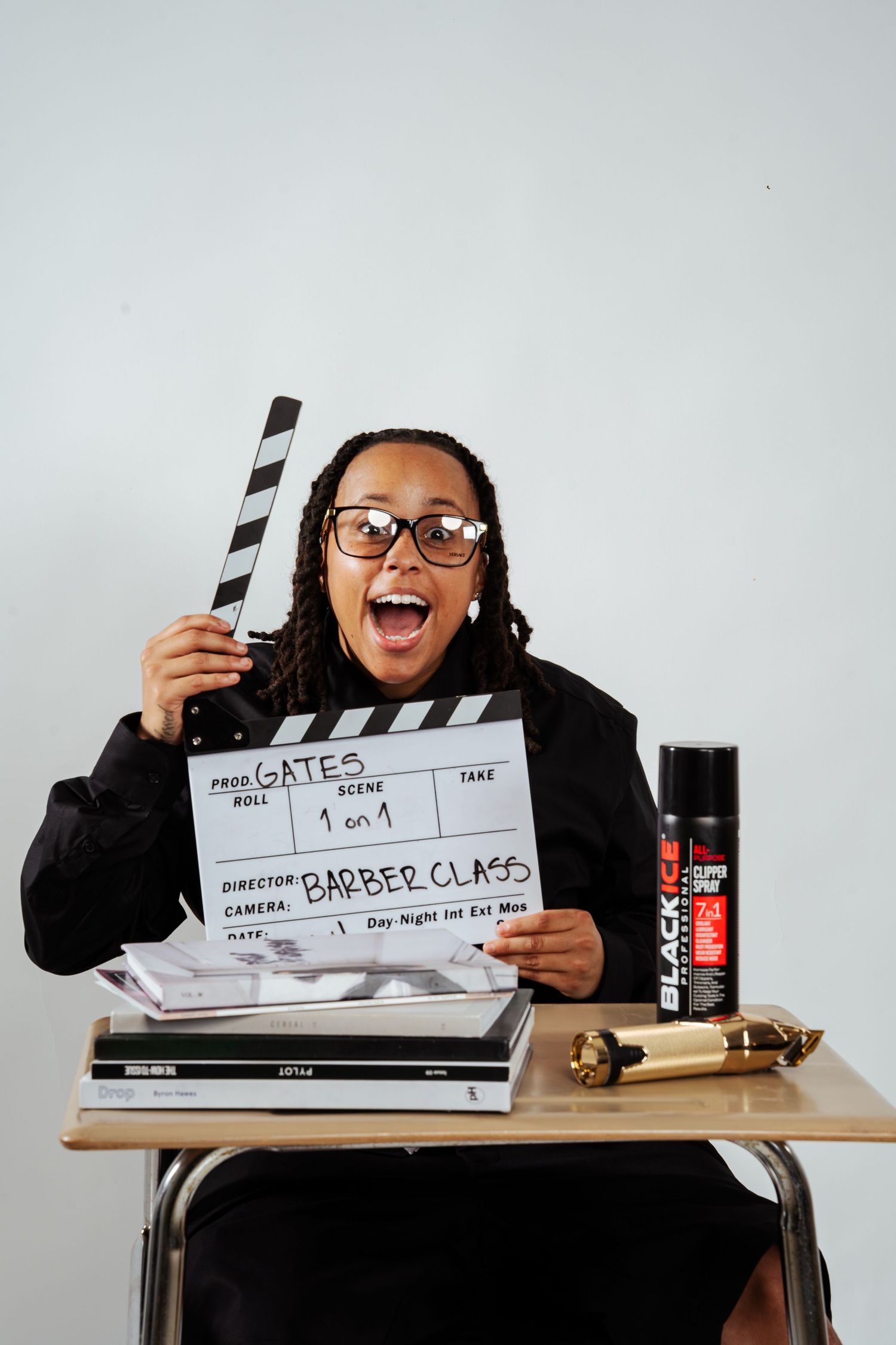 Excited person in black suit holding Barber Class film clapperboard with mouth open wide at school desk.