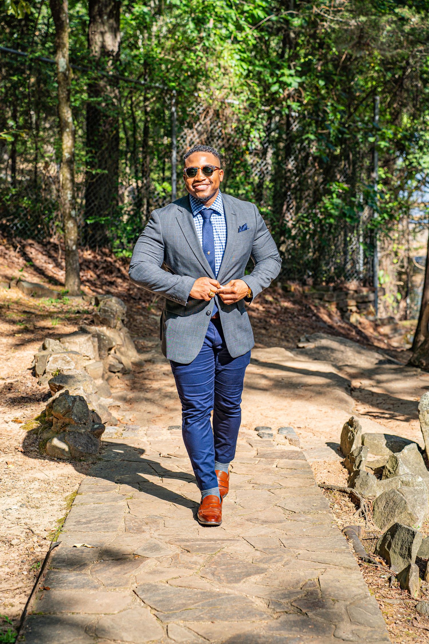 Stylish man in gray blazer, blue pants, and sunglasses walking on stone path in sunny forest.