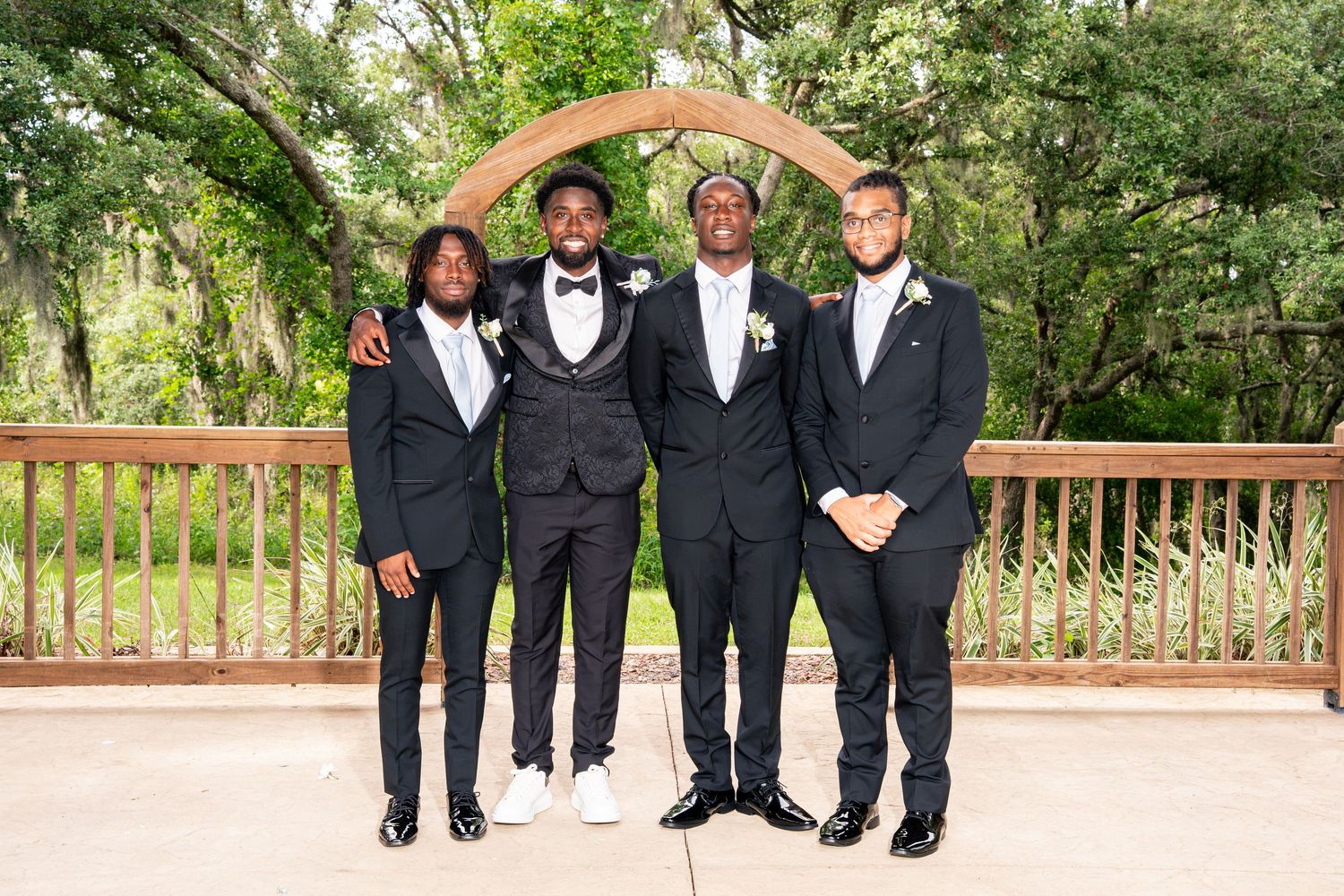Groom stands with three groomsmen in black tuxedos under wooden arch at wedding venue.