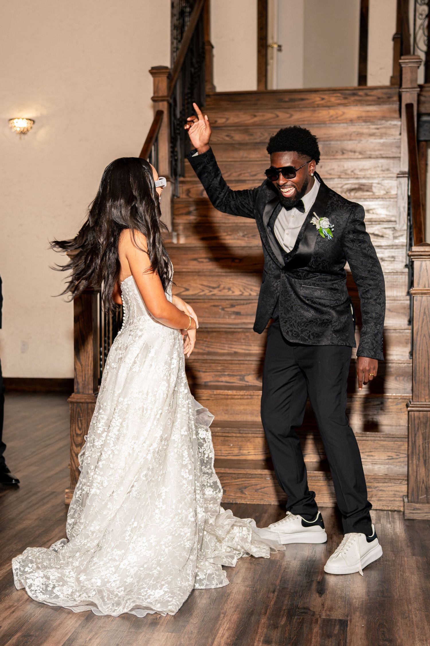 Bride and groom dance energetically near wooden staircase, groom in black tuxedo with sunglasses raising one hand.