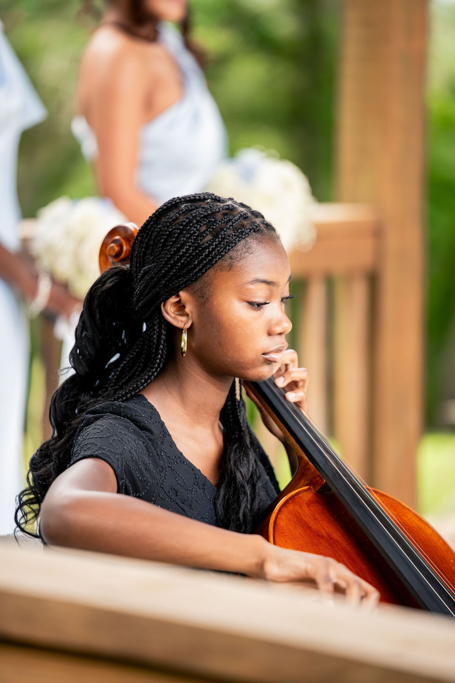 Young girl with braided hair plays cello at outdoor wedding ceremony.