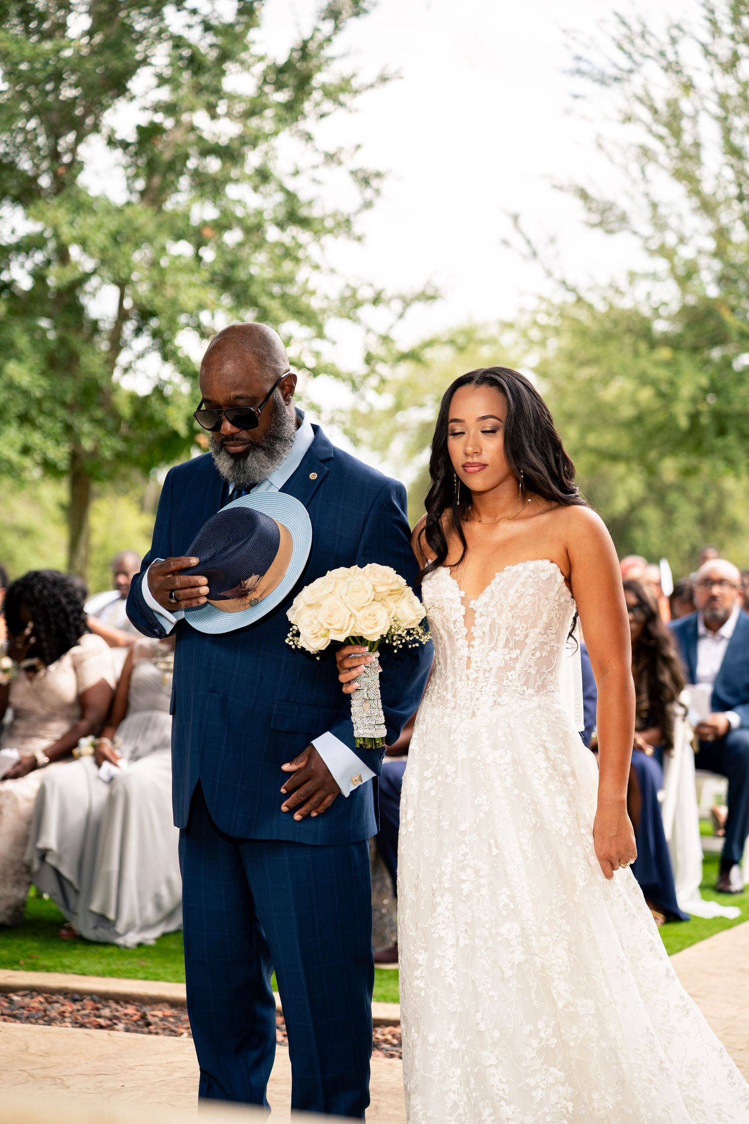 Father escorts bride down outdoor aisle, bride holding white bouquet in elegant lace wedding dress.