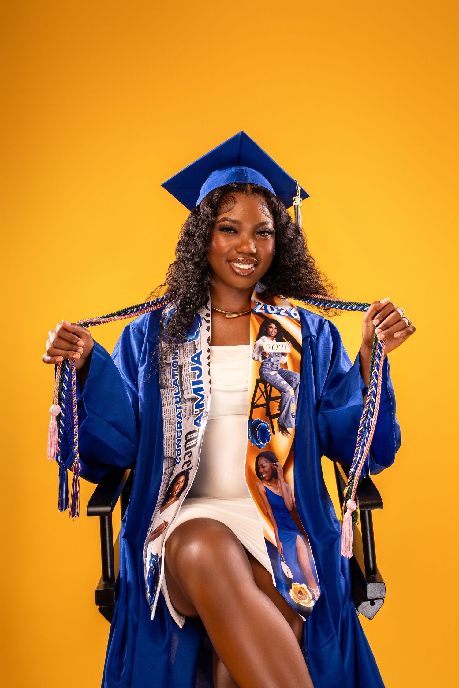Graduate in blue cap and gown sitting confidently holding honor cords on yellow background.