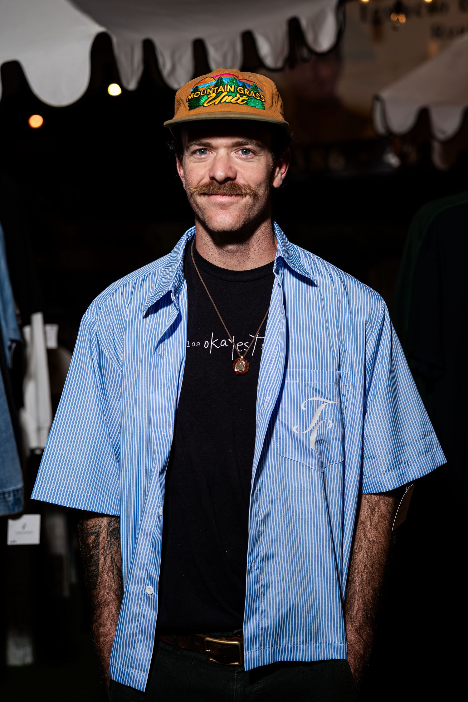 Smiling man with mustache wearing orange cap and blue striped Christian graphic shirt at event.