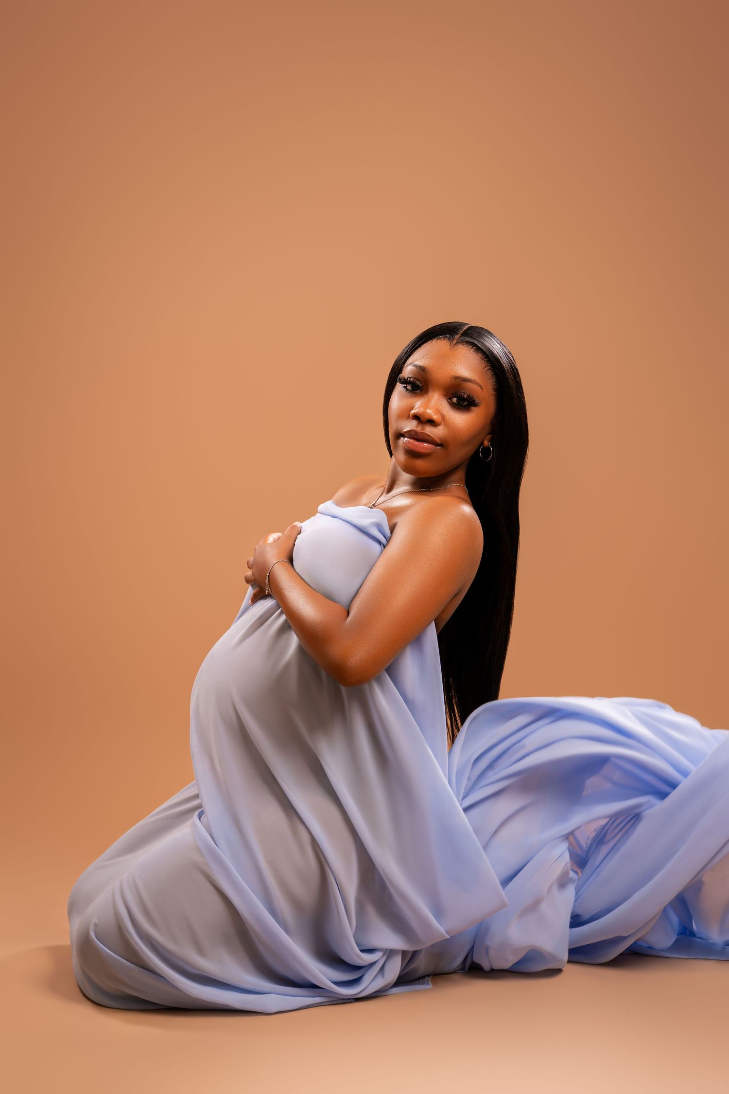 Pregnant woman in flowing light blue dress sitting gracefully on studio floor against warm beige background.