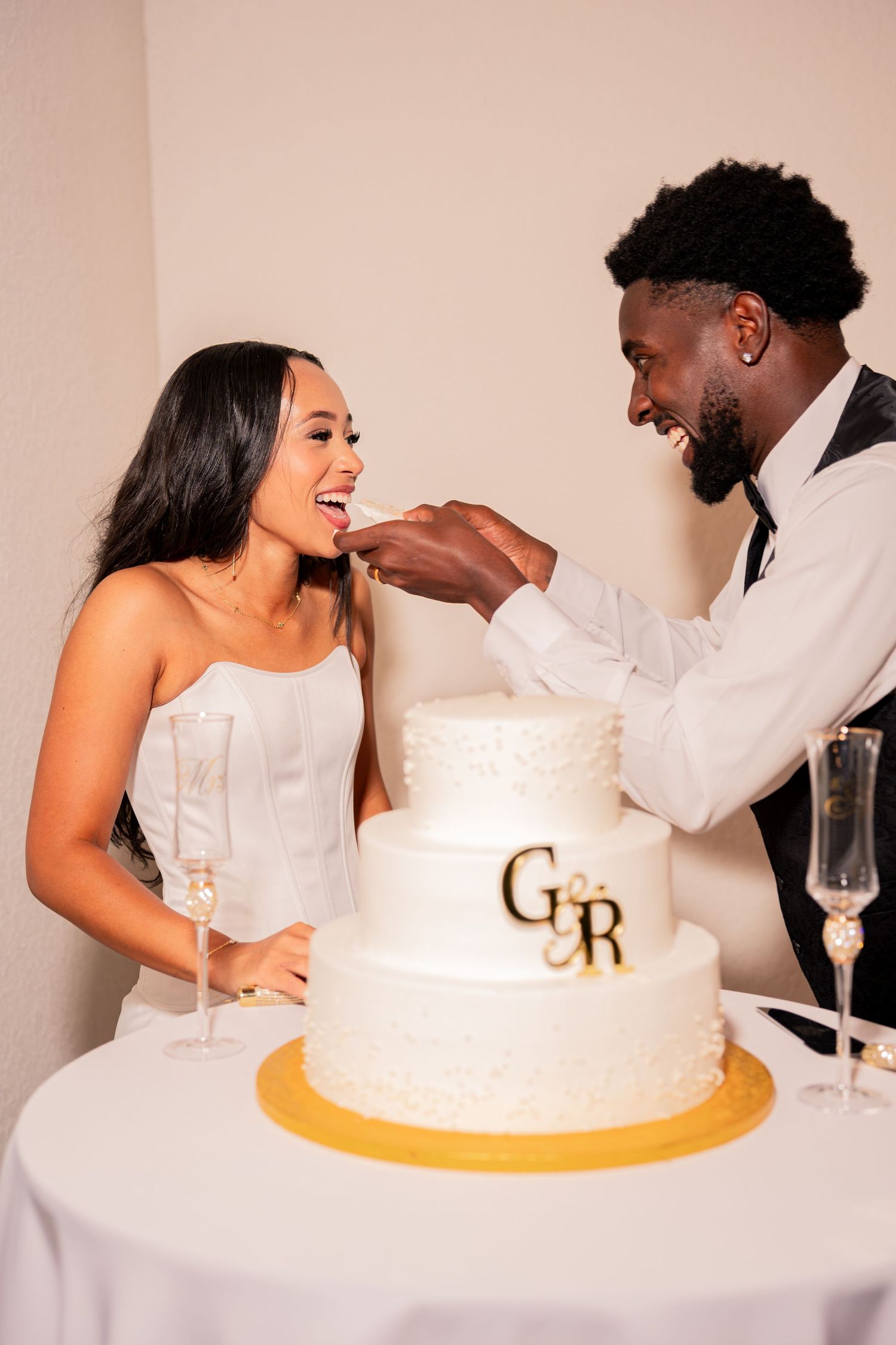 Groom feeding cake to laughing bride beside three-tier white wedding cake with gold monogram.