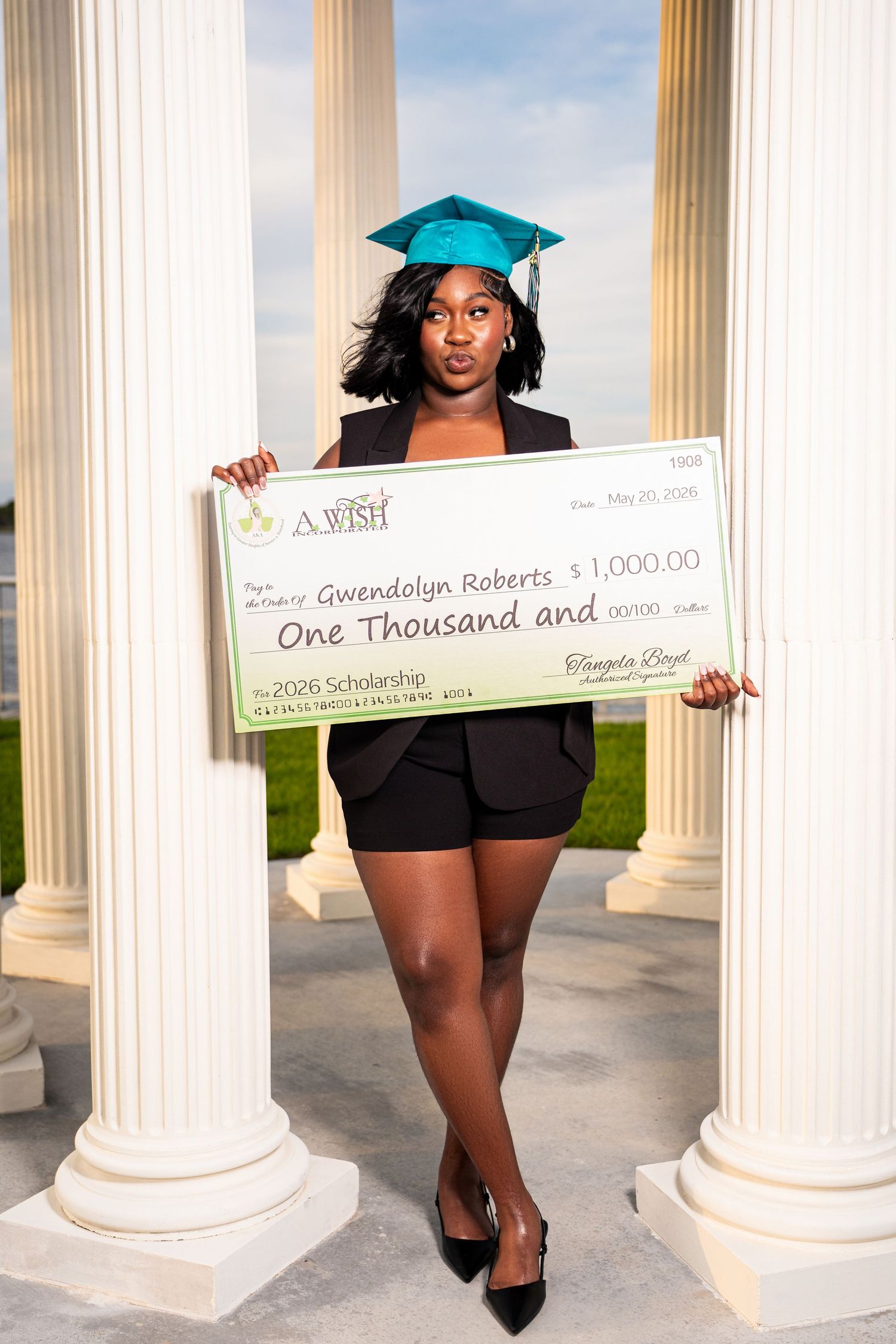 Black female graduate in teal cap holding large $1,000 scholarship check between white columns at sunset.