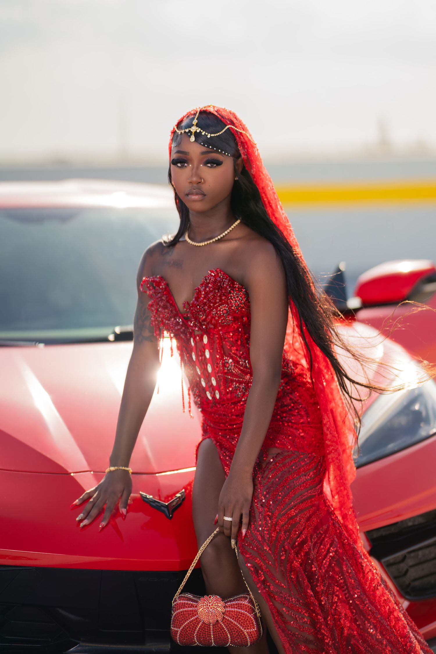A stunning Black woman in a red beaded gown and headdress poses beside a red Corvette sports car.
