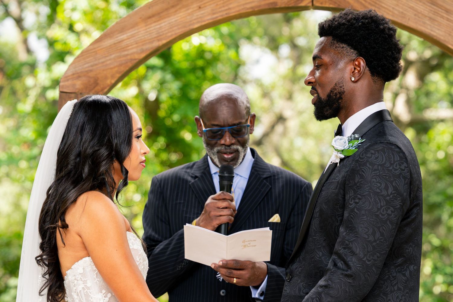 Officiant reads vows as bride and groom stand under wooden arch during outdoor wedding ceremony.