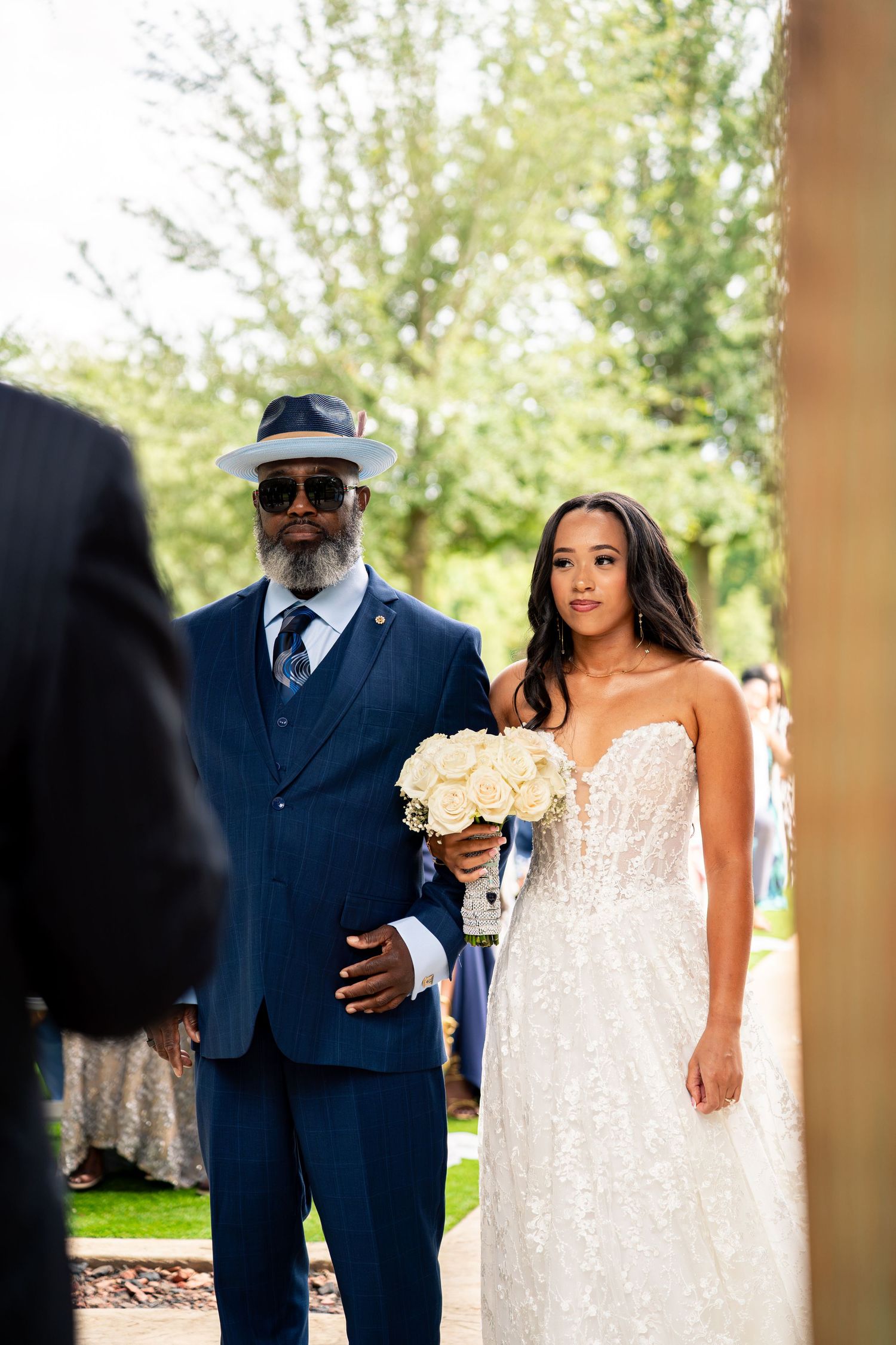 Bride in strapless lace gown holds white bouquet while being walked down aisle by father in blue suit.