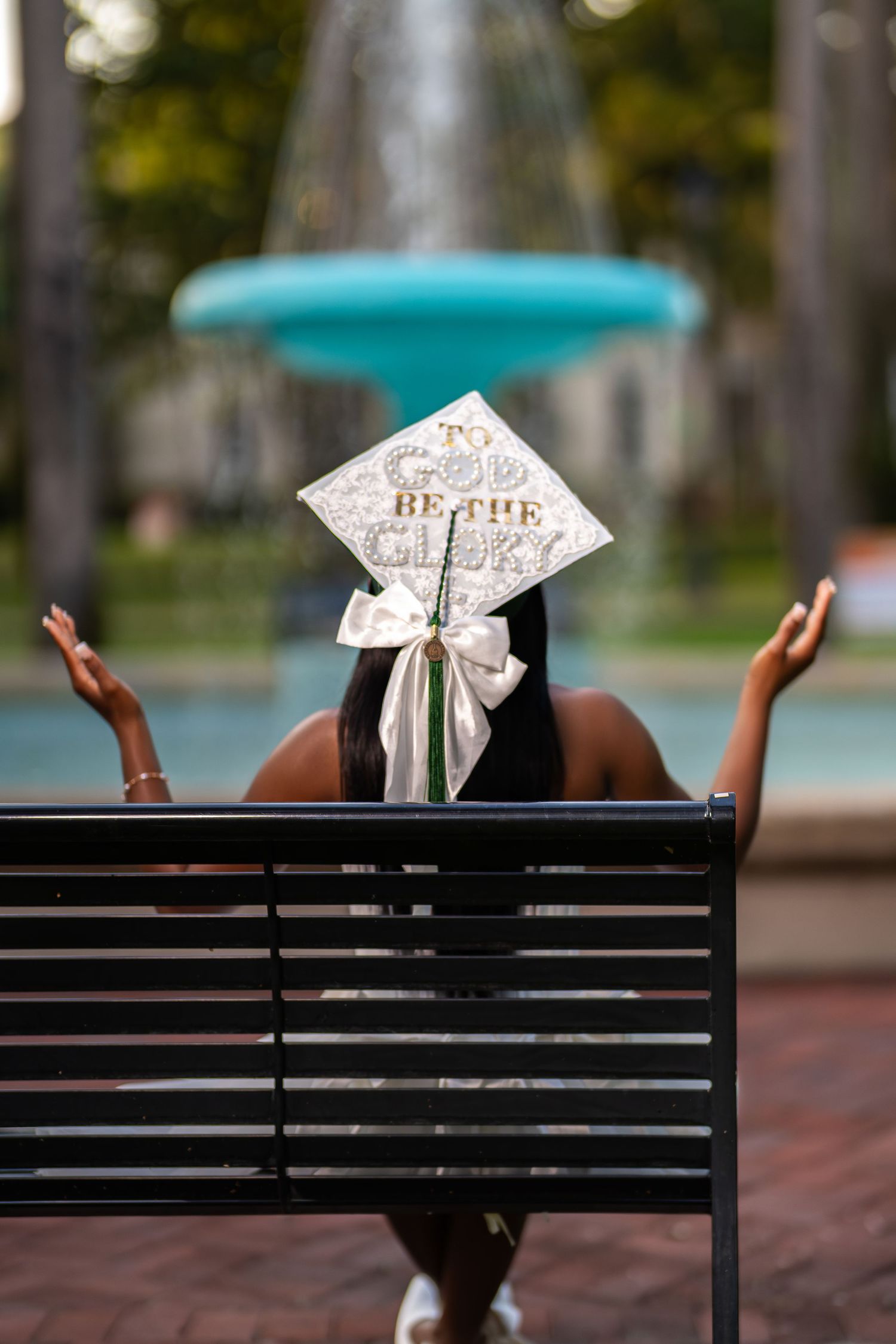 Graduate in decorated cap reading To God Be The Glory raises hands near fountain on campus bench.