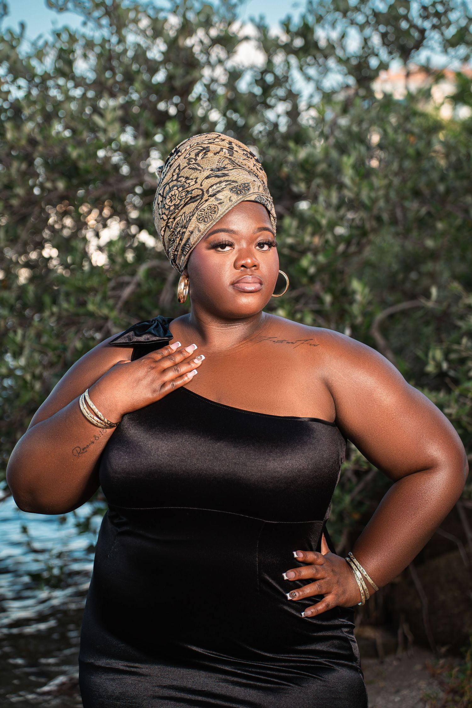 Confident plus-size Black woman in black one-shoulder dress and gold headwrap poses near trees.