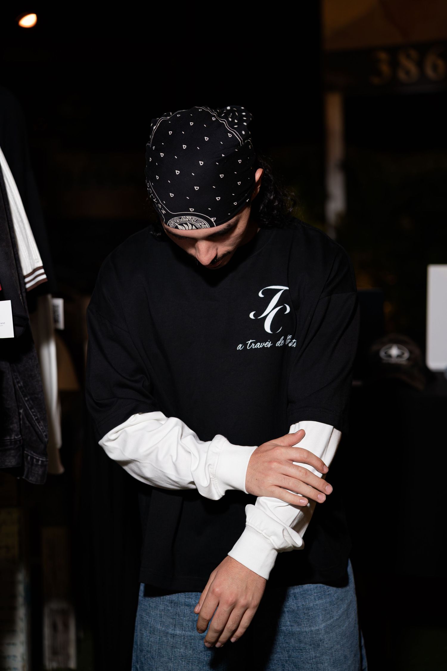 Man in black TC logo sweatshirt and bandana cap looking down at outdoor night market.