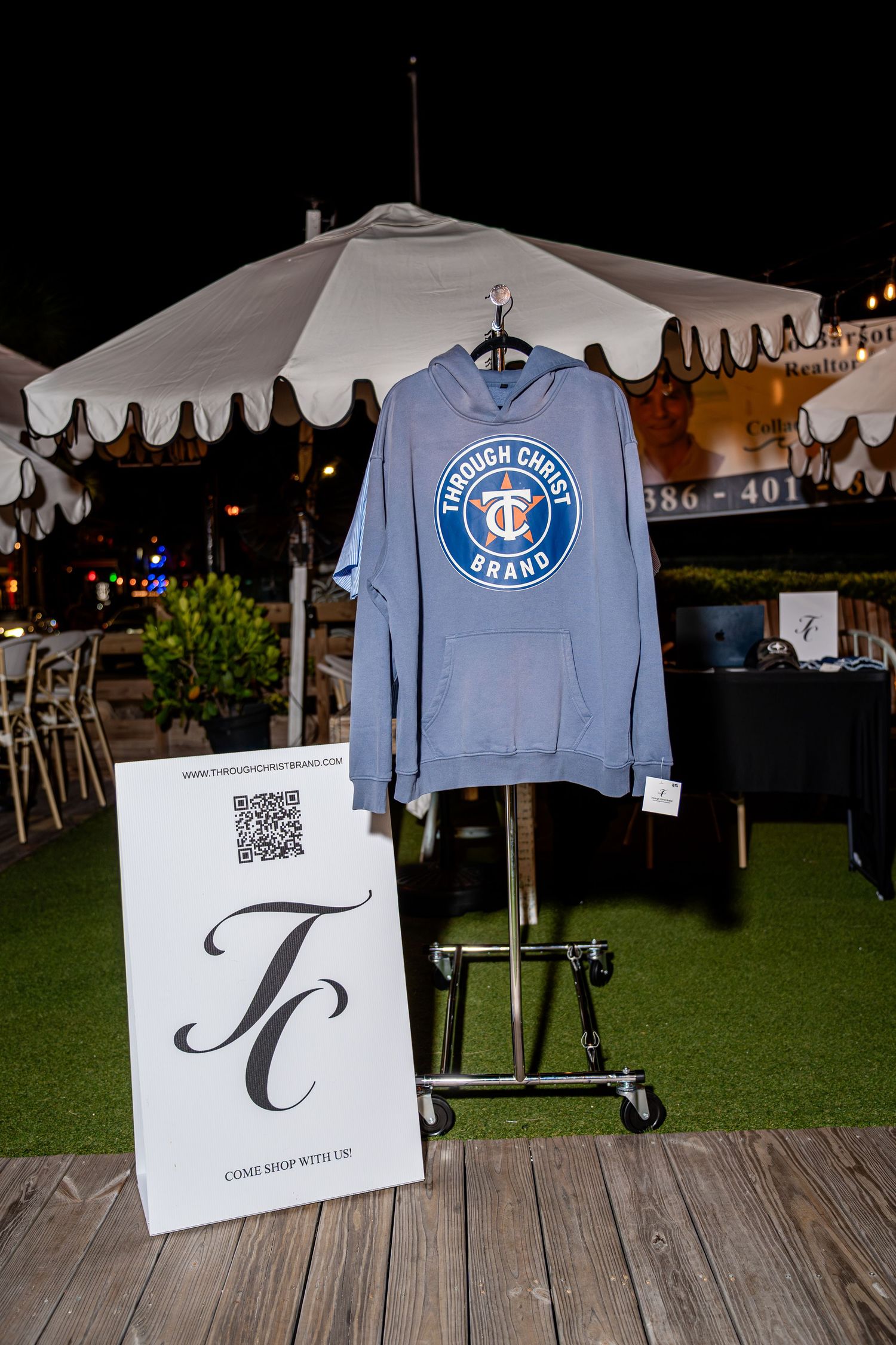 Blue Through Christ Brand hoodie displayed on rack with TC logo sign at outdoor night event.
