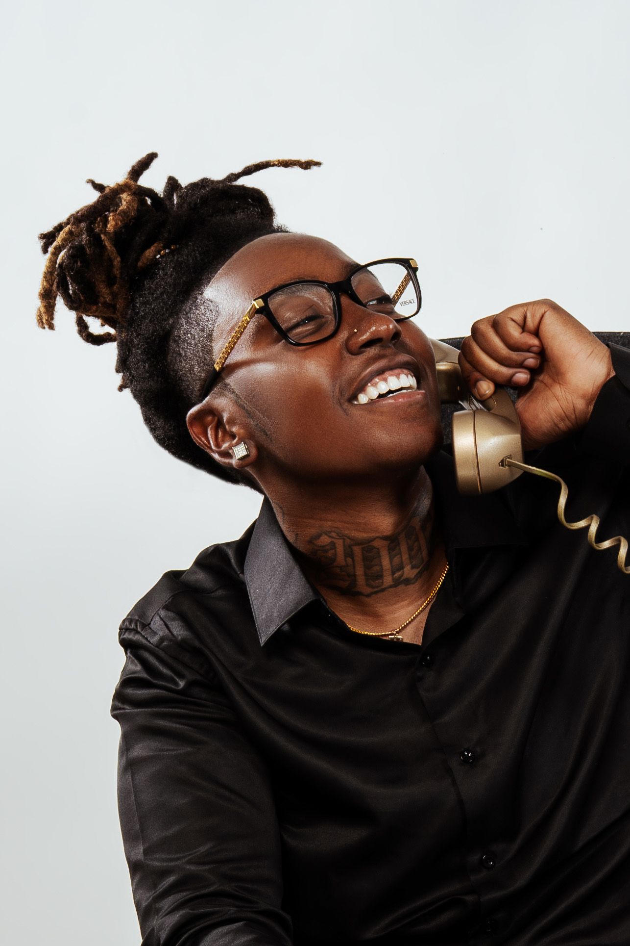 Laughing young Black man with dreadlocks and glasses holding a gold vintage telephone against light background.