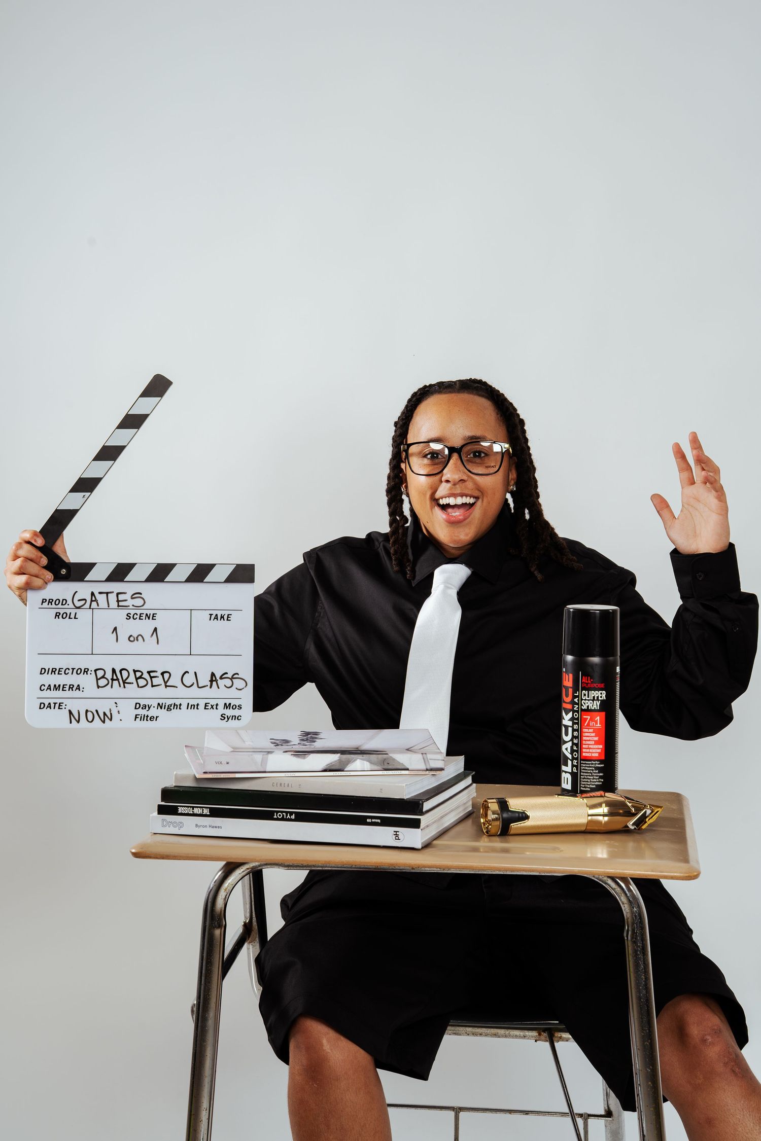 Excited person in black suit holding film clapperboard labeled Barber Class at school desk with Black Ice spray can.
