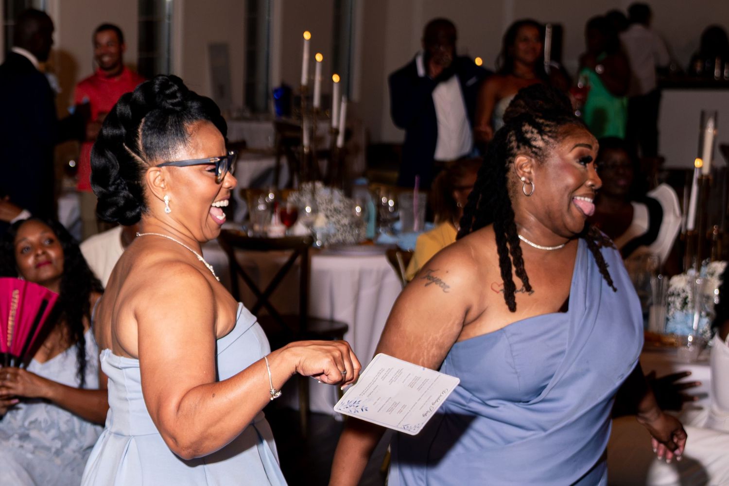 Two women in blue dresses laugh and dance together at wedding reception celebration.