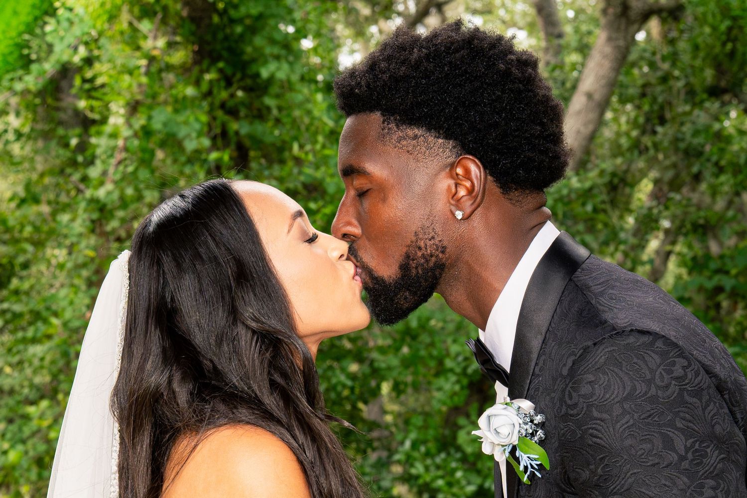 Bride and groom sharing a romantic first kiss surrounded by lush green foliage at their outdoor wedding.