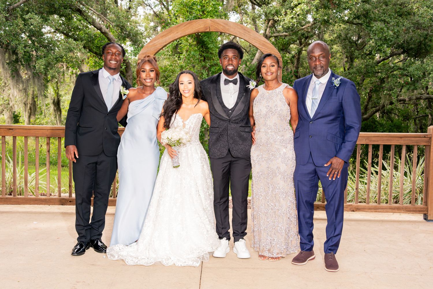 Wedding party posing under wooden arch at outdoor ceremony with bride and groom.