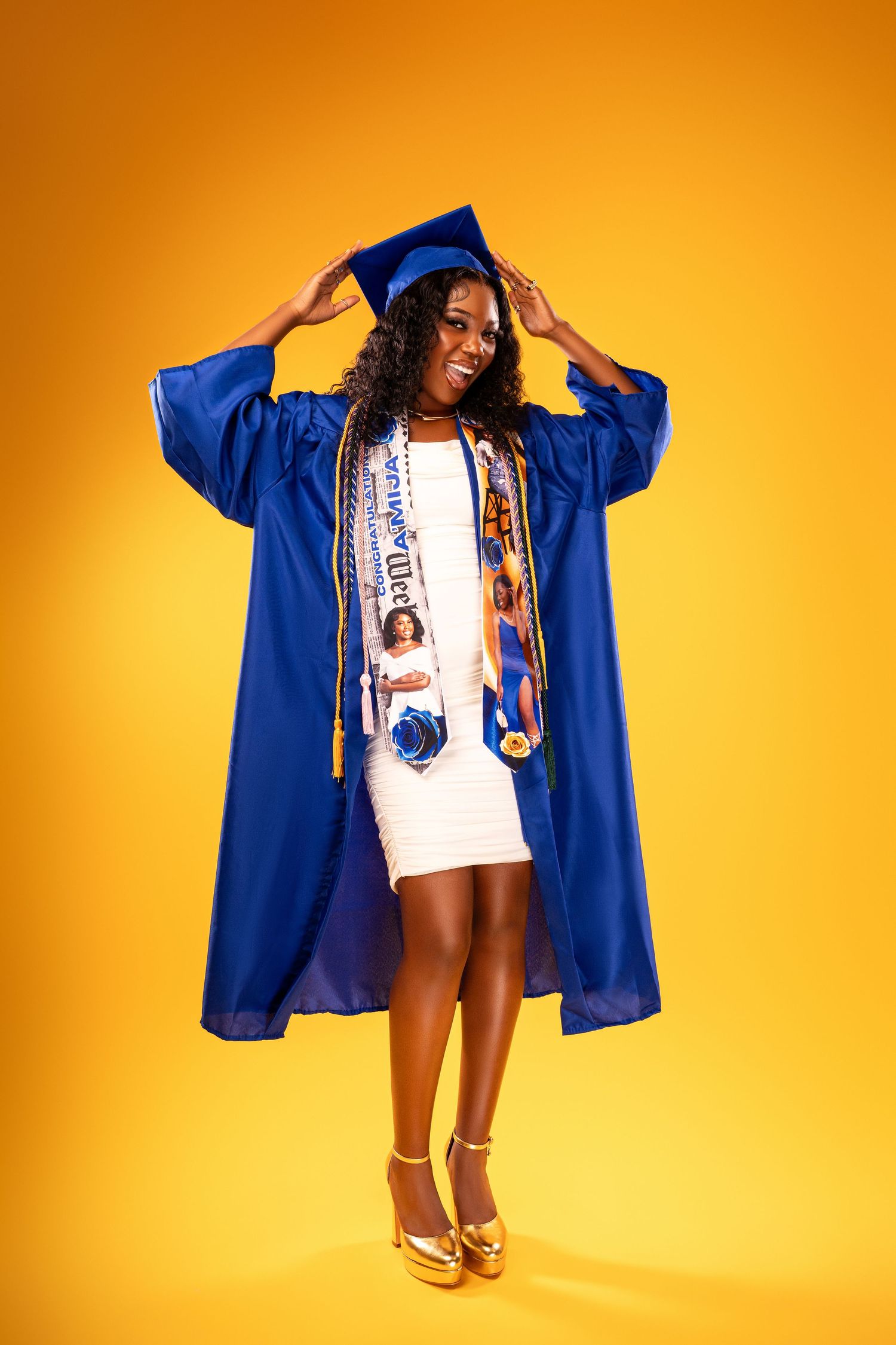 Full-length portrait of graduate in blue cap and gown with sash on yellow background.
