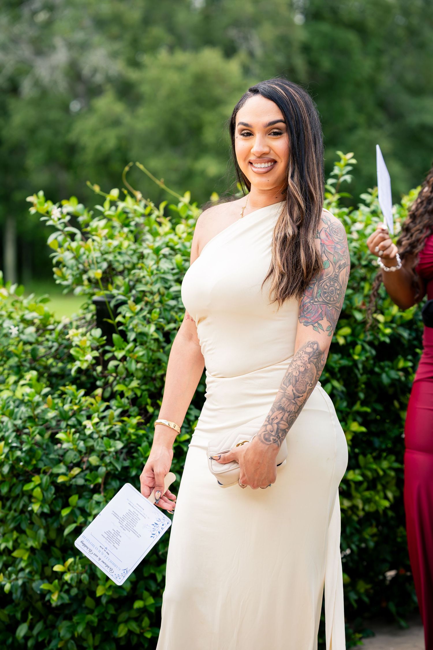 Woman in cream one-shoulder dress with tattoo sleeve smiling outdoors at a wedding event.