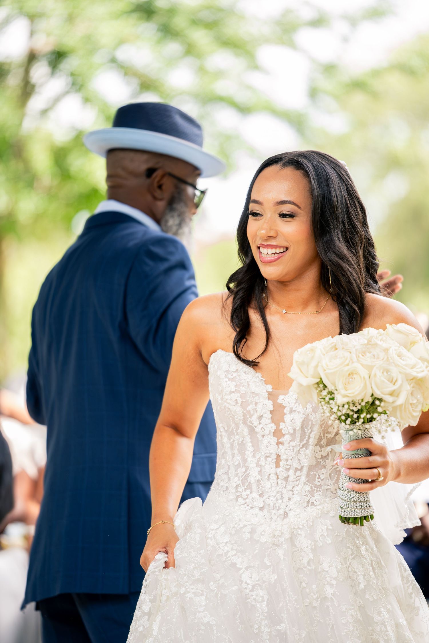 Smiling bride in lace gown holds white rose bouquet while walking down aisle with older man in navy suit and hat.