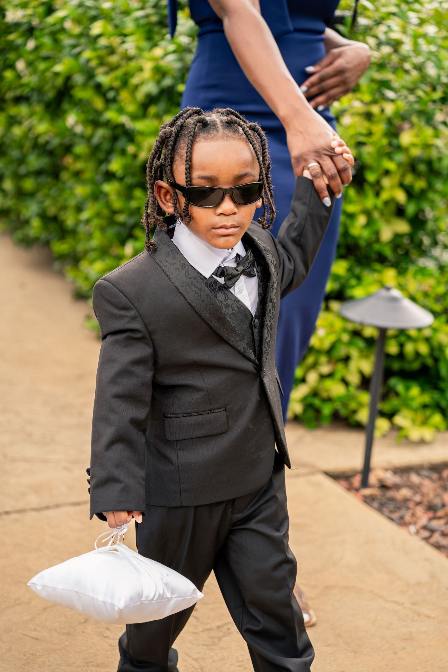 Stylish young ring bearer in black tuxedo and sunglasses carries white pillow down the wedding ceremony aisle.