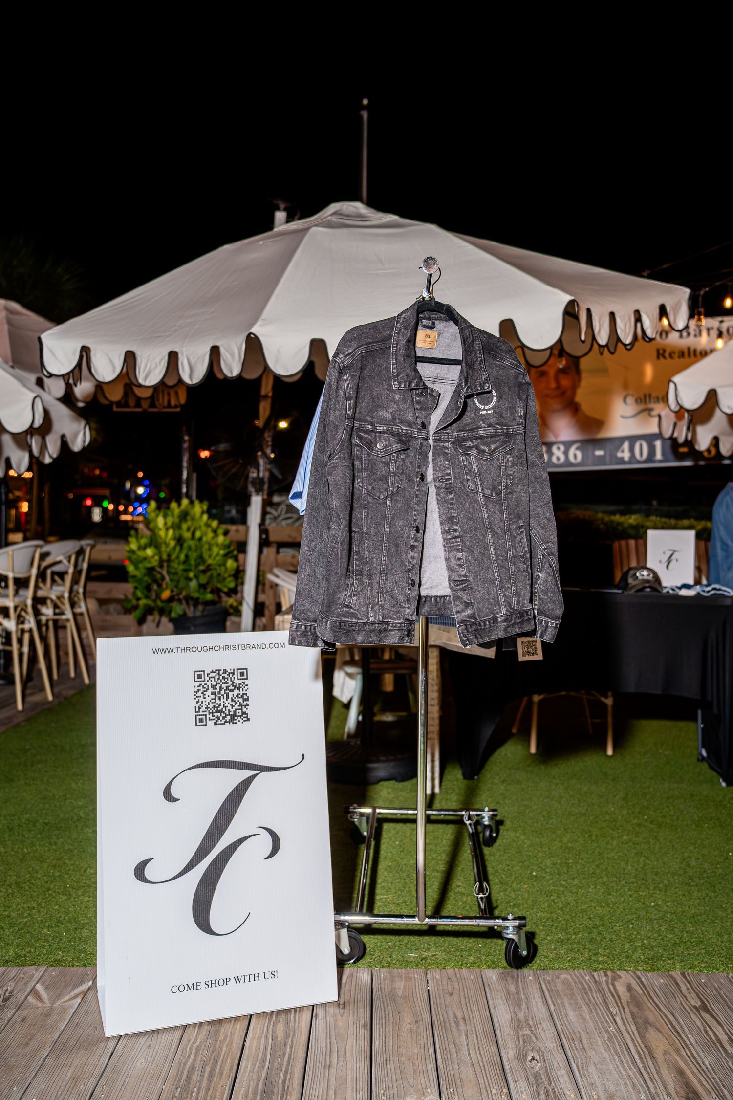 Dark gray denim jacket displayed on metal rack beside TC brand QR code sign at outdoor event.