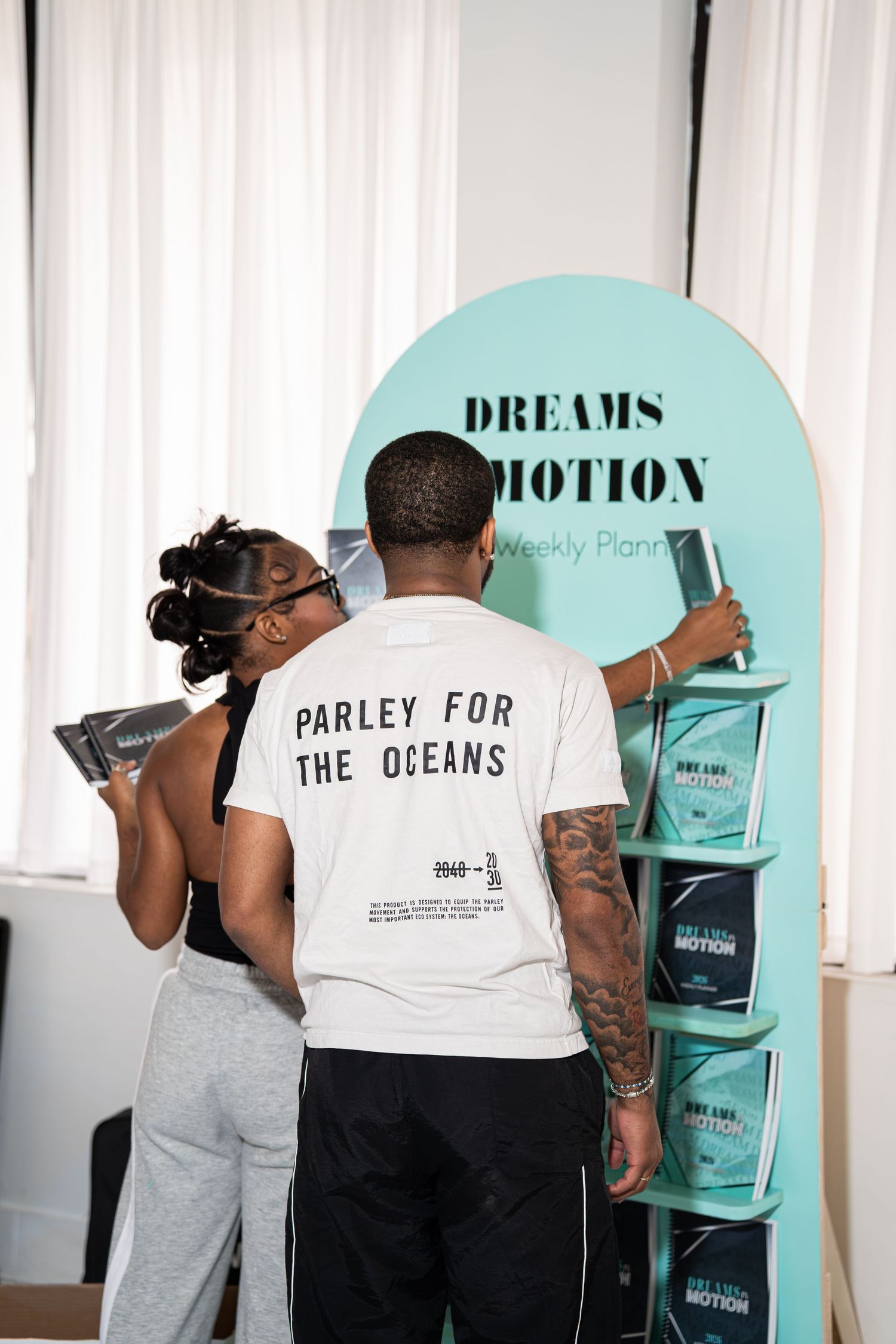 Two attendees browse Dreams in Motion weekly planners at a teal branded display booth.
