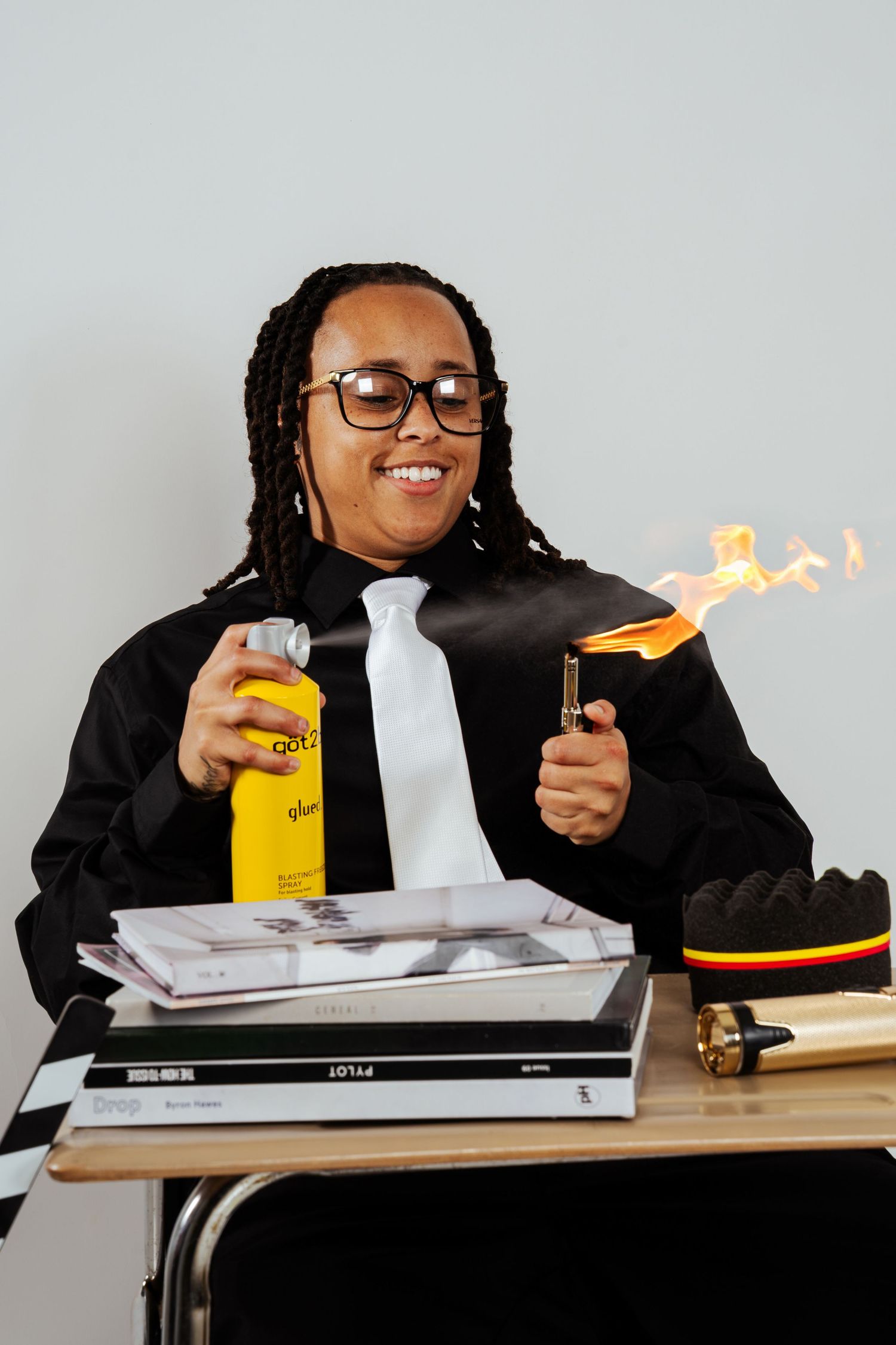 Smiling person in black suit creating fire with lighter and spray can while seated at school desk with books.