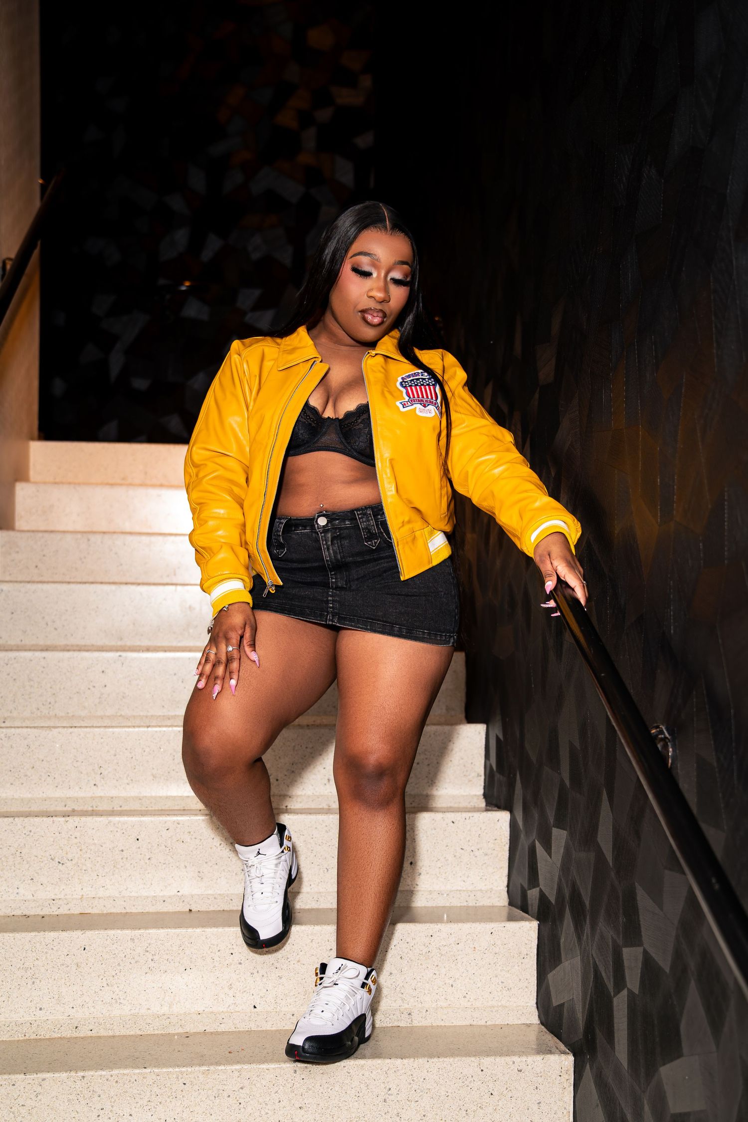 Black woman in yellow bomber jacket, black mini skirt, and white Jordans poses on indoor staircase.