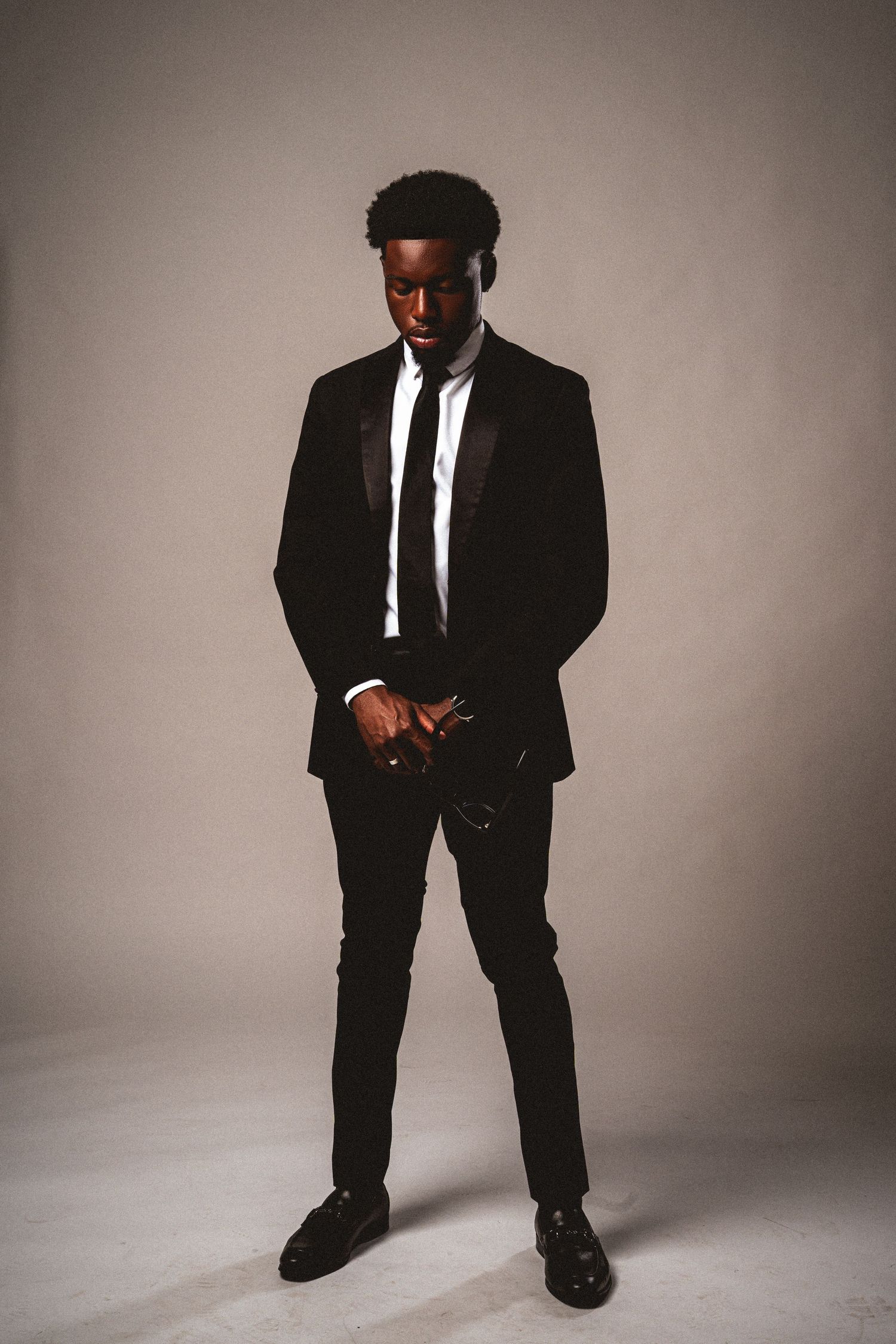A stylish Black man in a black tuxedo stands with clasped hands against a neutral gray backdrop.