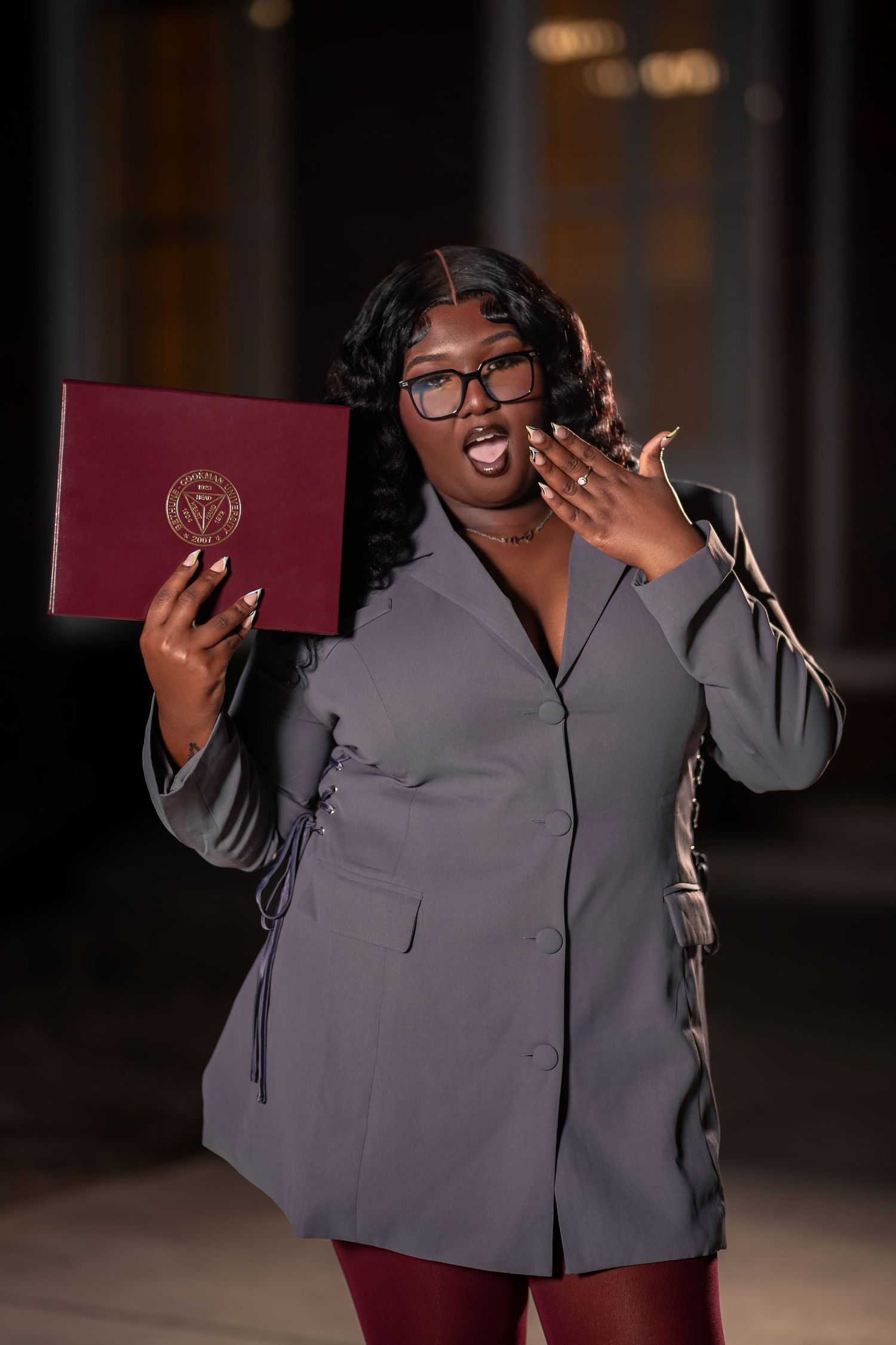 Excited graduate in gray blazer holds maroon diploma cover while blowing a celebratory kiss to camera.