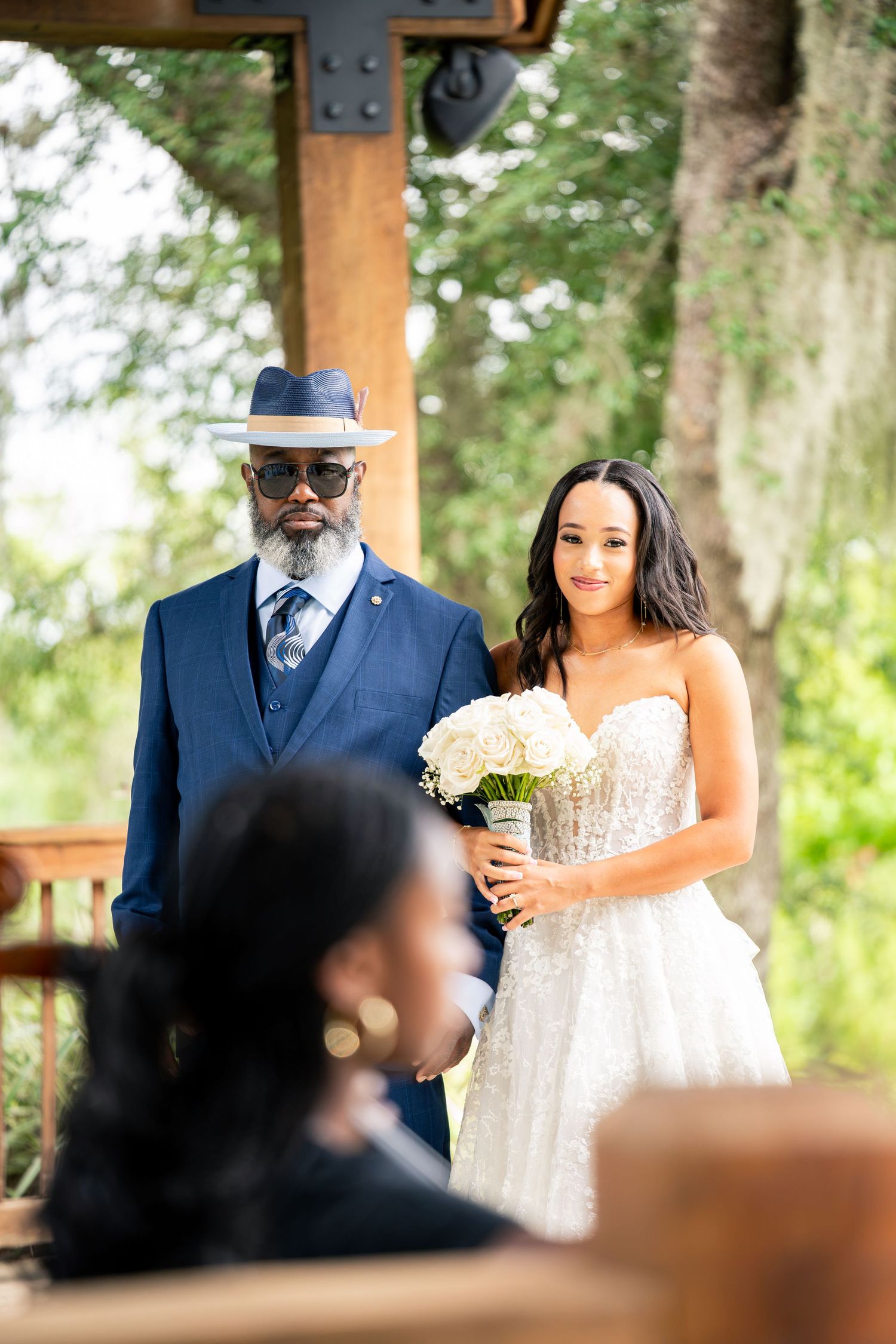 Bride in lace gown holds white bouquet beside elegantly dressed man in navy suit and hat at outdoor wedding ceremony.