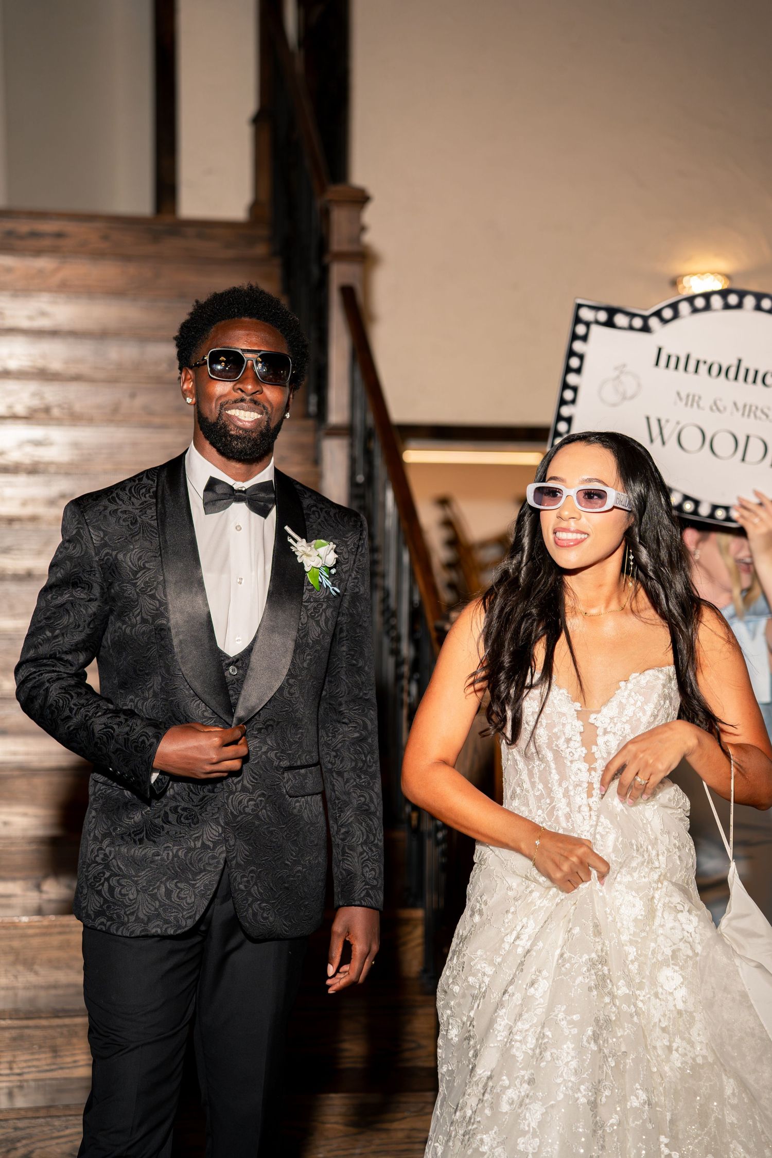 Newlyweds make grand reception entrance wearing sunglasses, groom in black patterned tuxedo, bride in lace wedding gown.