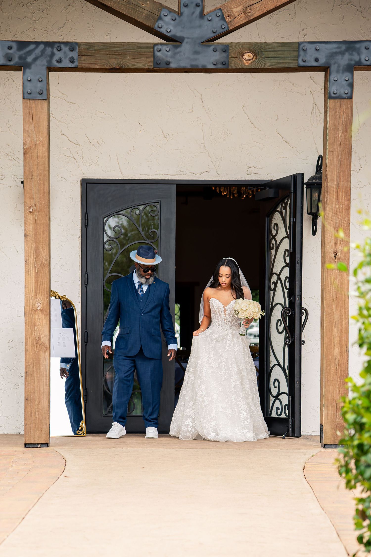 Bride in lace ballgown and veil exits venue doors with escort in navy suit and hat for ceremony processional.