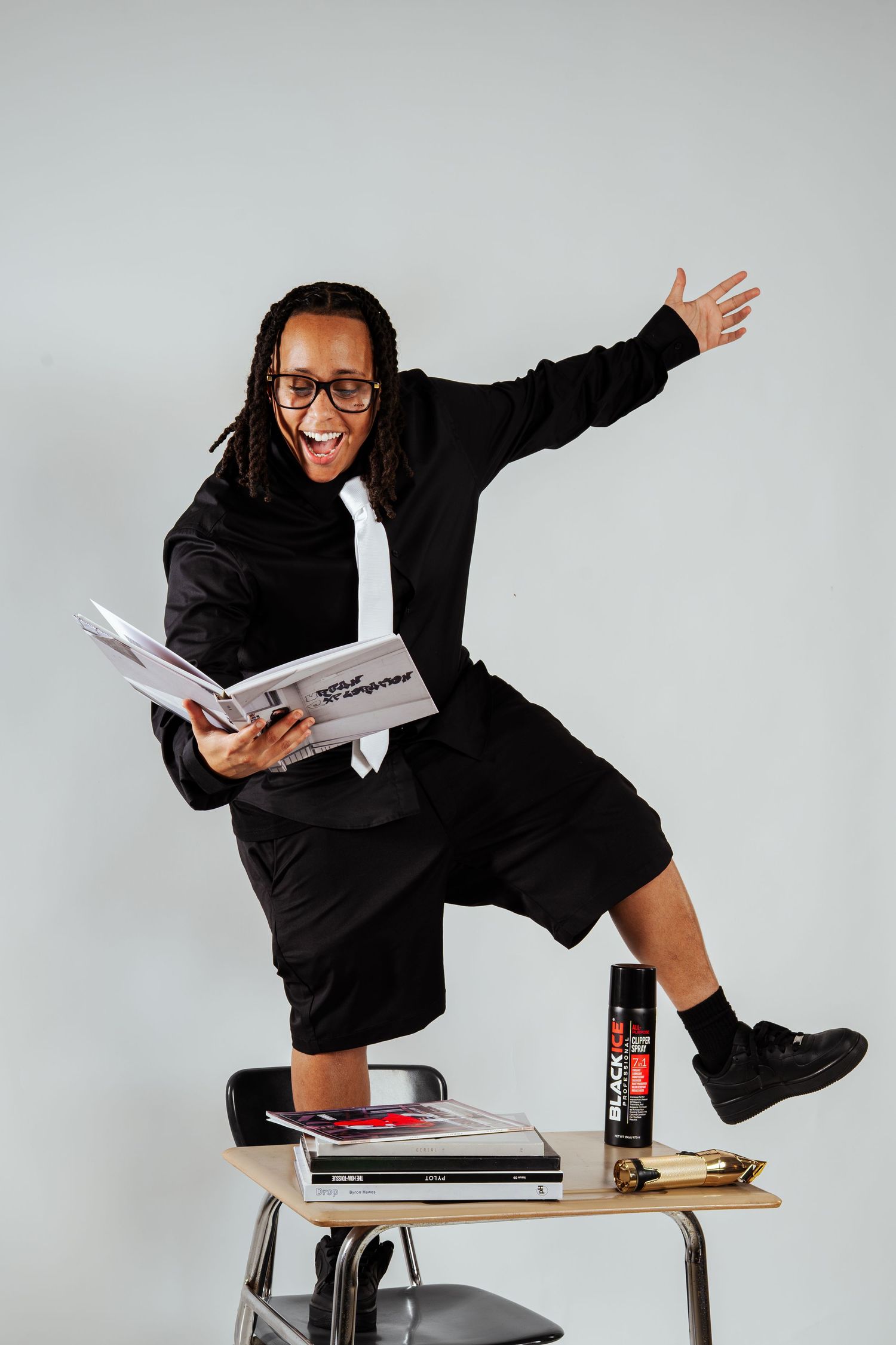 Energetic person in black suit standing on school desk holding open book with Black Ice spray can nearby.