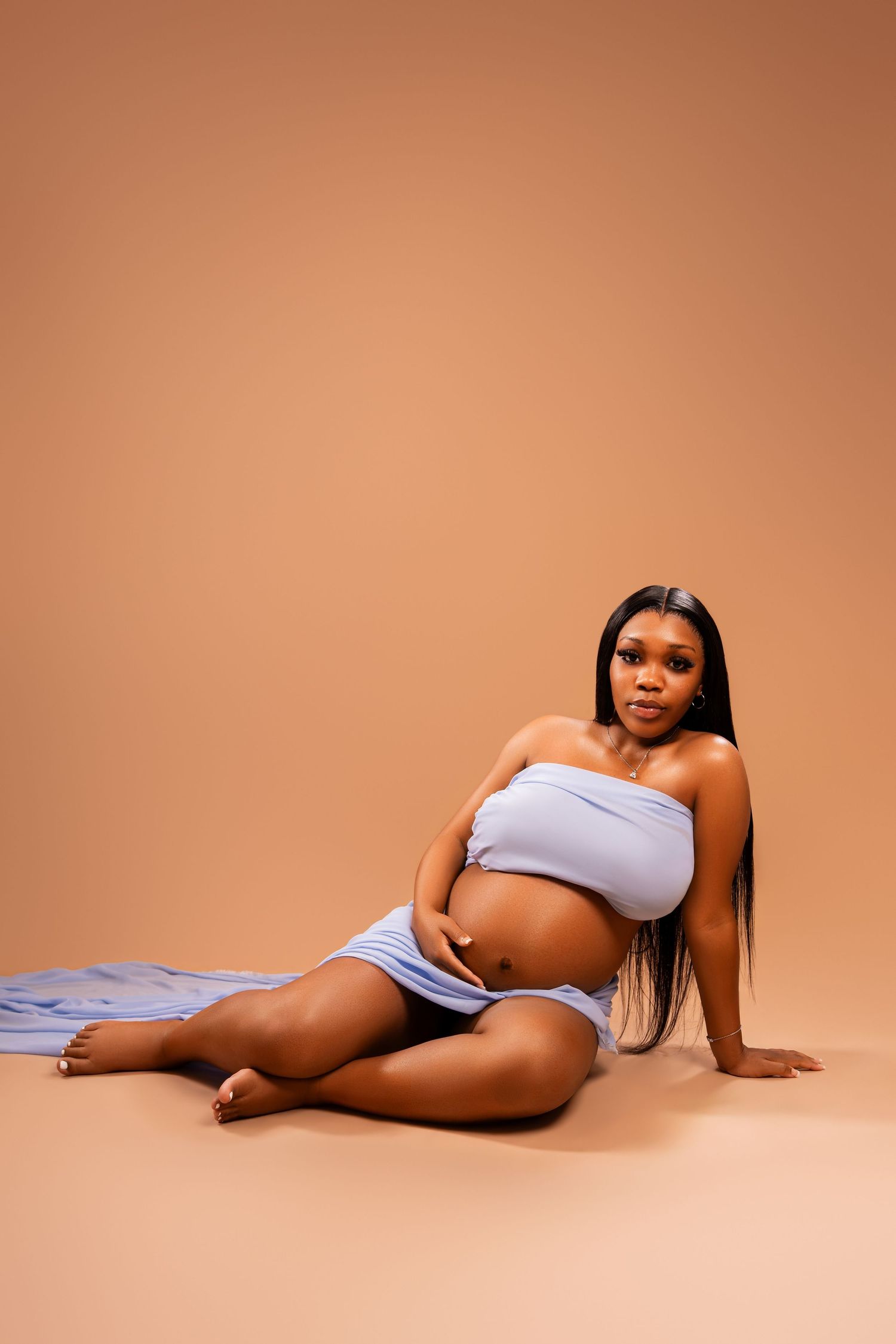 Pregnant Black woman in blue bandeau top sitting gracefully on studio floor against warm beige background.