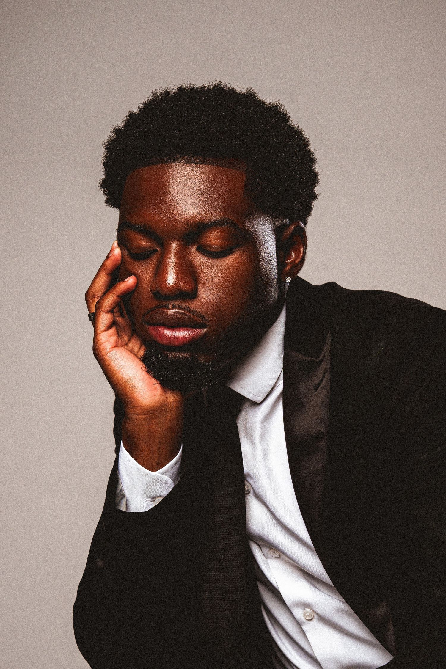 Pensive Black man in black suit with eyes closed, hand on face.