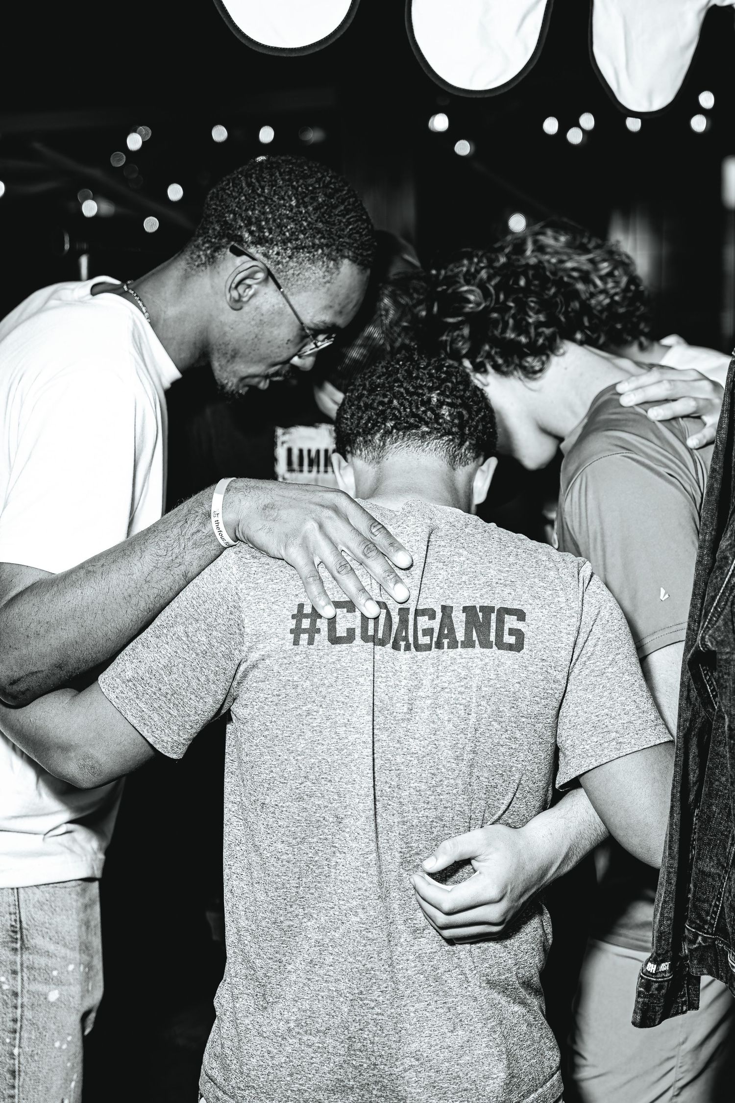 Black and white photo of three young men praying together wearing #CUDAGANG shirts.