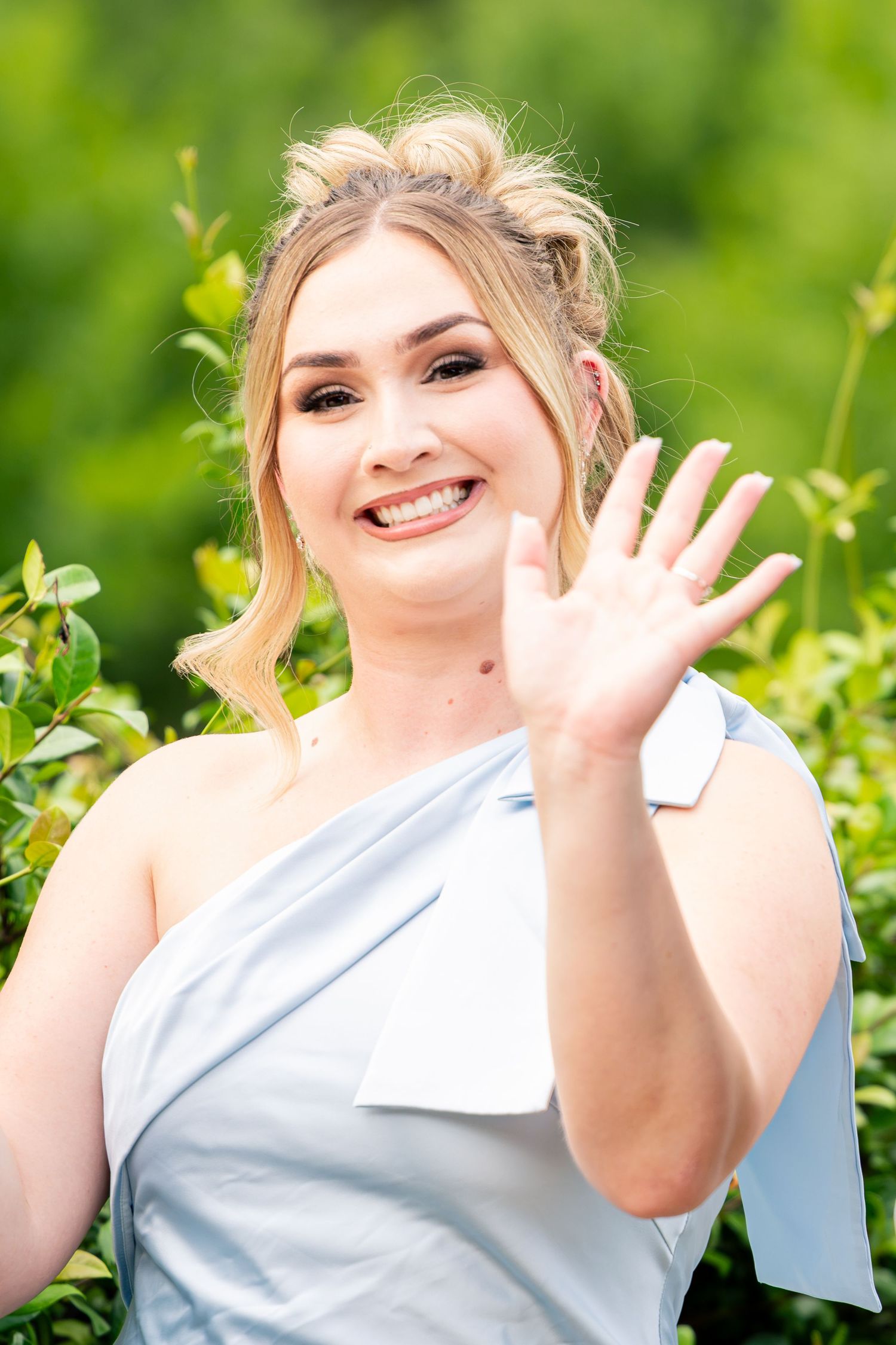 Smiling blonde woman in light blue one-shoulder dress waving at an outdoor wedding.
