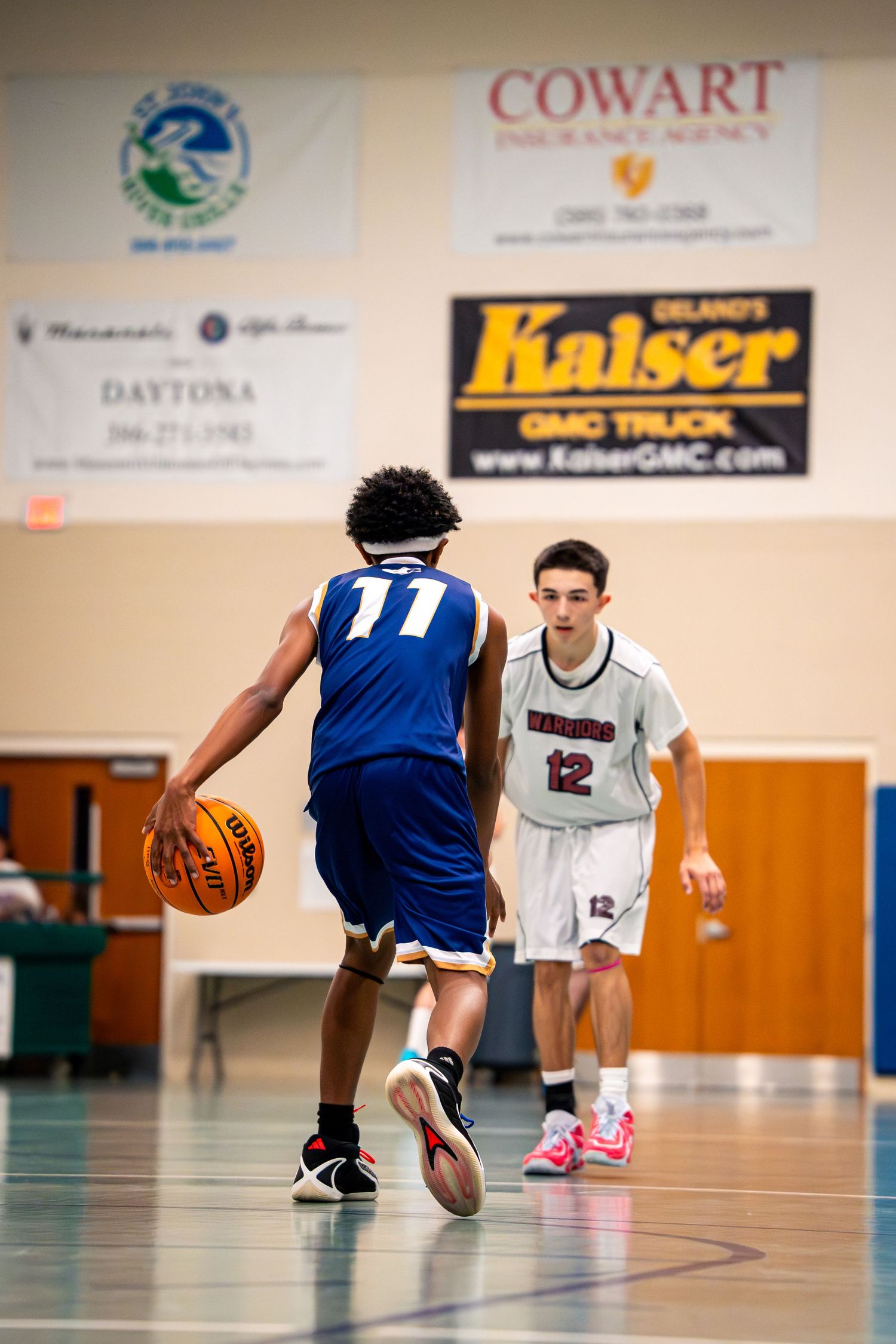 Player #11 in blue dribbles basketball while #12 in white defends during indoor youth basketball game.
